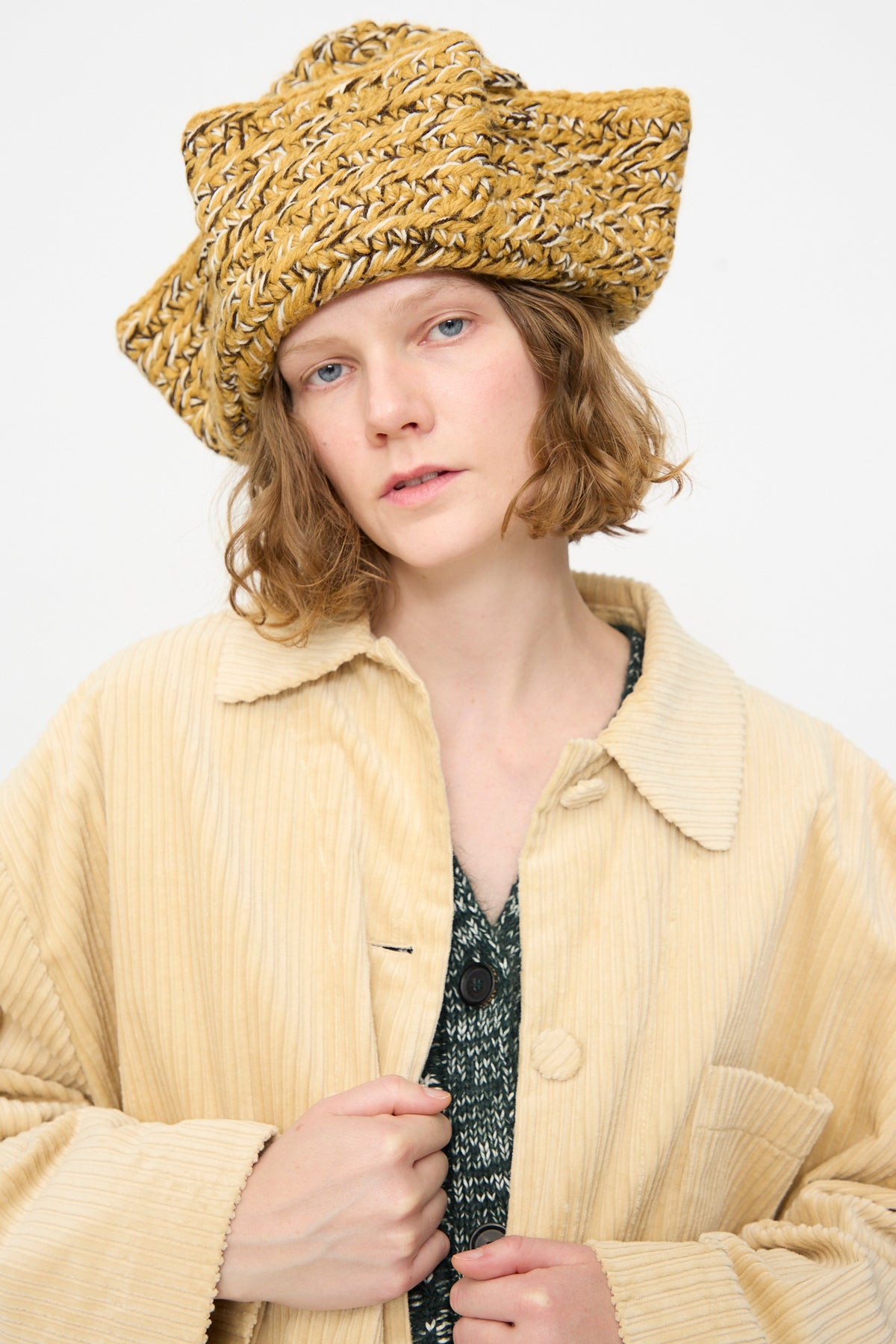 Wearing the Tricorn in Dark Gold Marle by Rachel Comey—a textured yellow triangular hat crafted from Andean Highland wool—paired with a light beige corduroy coat and a green patterned top, the person stands against a plain white background.