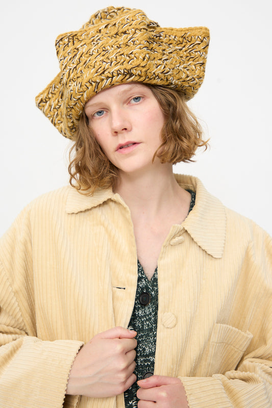 Wearing the Tricorn in Dark Gold Marle by Rachel Comey—a textured yellow triangular hat crafted from Andean Highland wool—paired with a light beige corduroy coat and a green patterned top, the person stands against a plain white background.