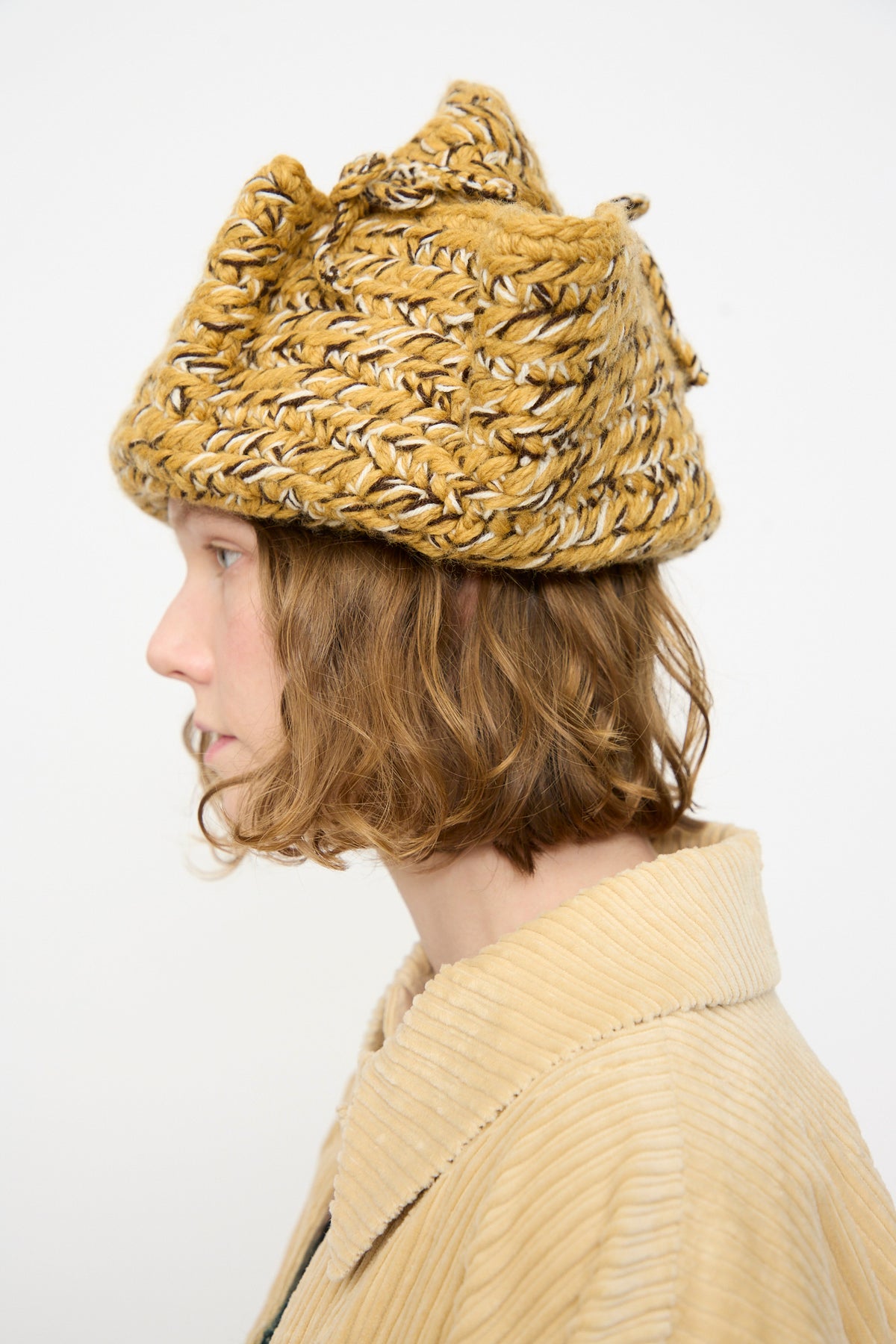 A person with light brown, wavy hair wears the Rachel Comey Tricorn hat in Dark Gold Marle—a textured, yellow knit triangular hat crafted from Andean Highland wool—and a beige corduroy jacket, shown in profile against a plain white background.