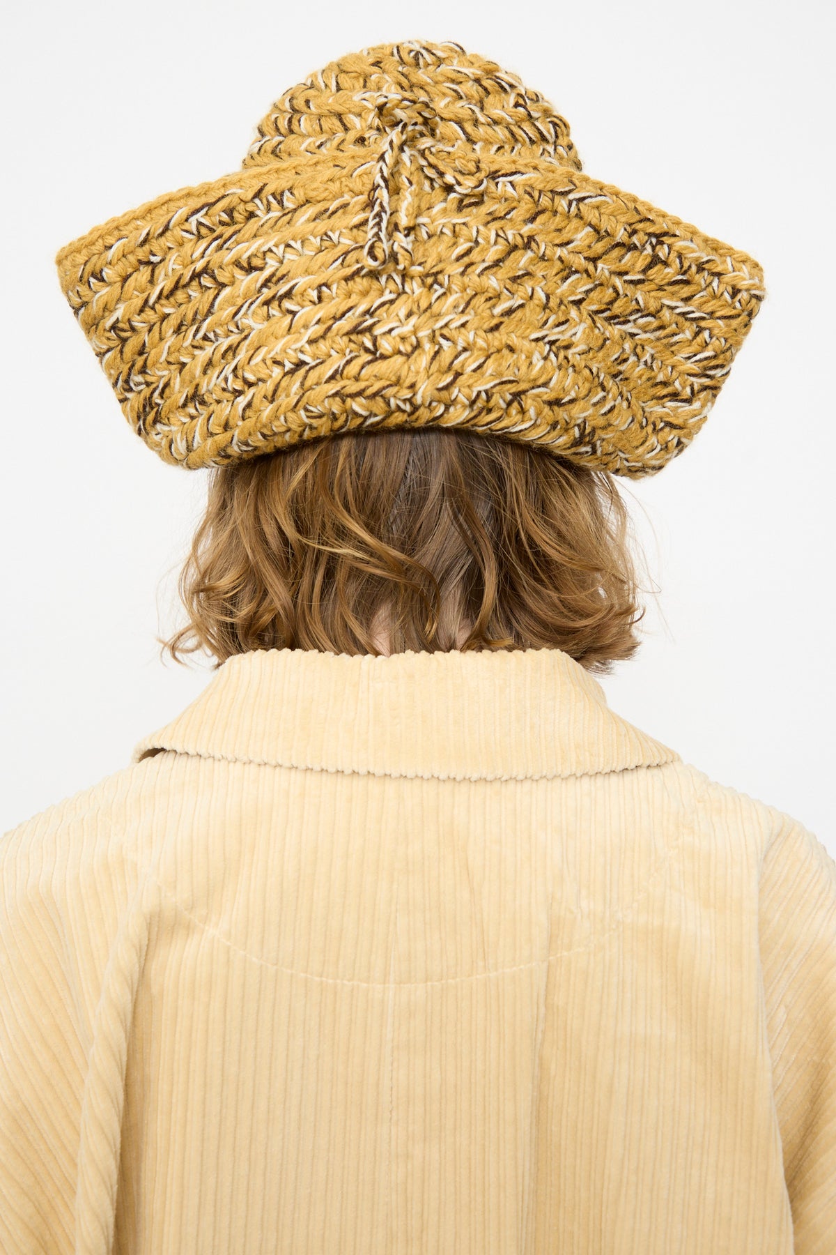 A person with shoulder-length hair, seen from behind against a plain white background, wears a tan corduroy jacket and the Rachel Comey Tricorn in Dark Gold Marle—a wide, woven straw hat featuring subtle knit accents inspired by Andean Highland wool craftsmanship.