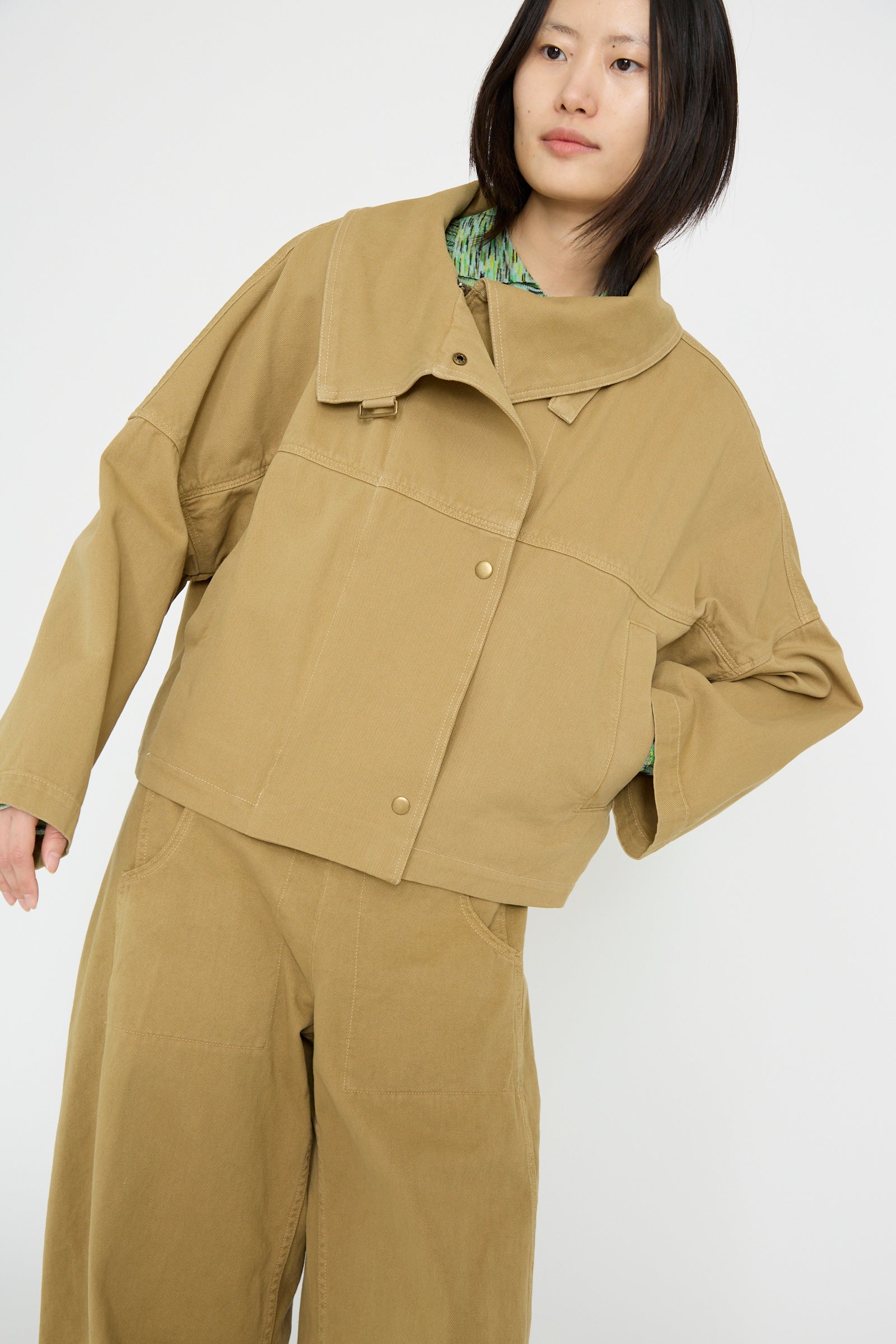 Against a plain white background, a person is in the oversized Vansire Jacket by Rachel Comey in Olive, with matching pants and a hand in their pocket.