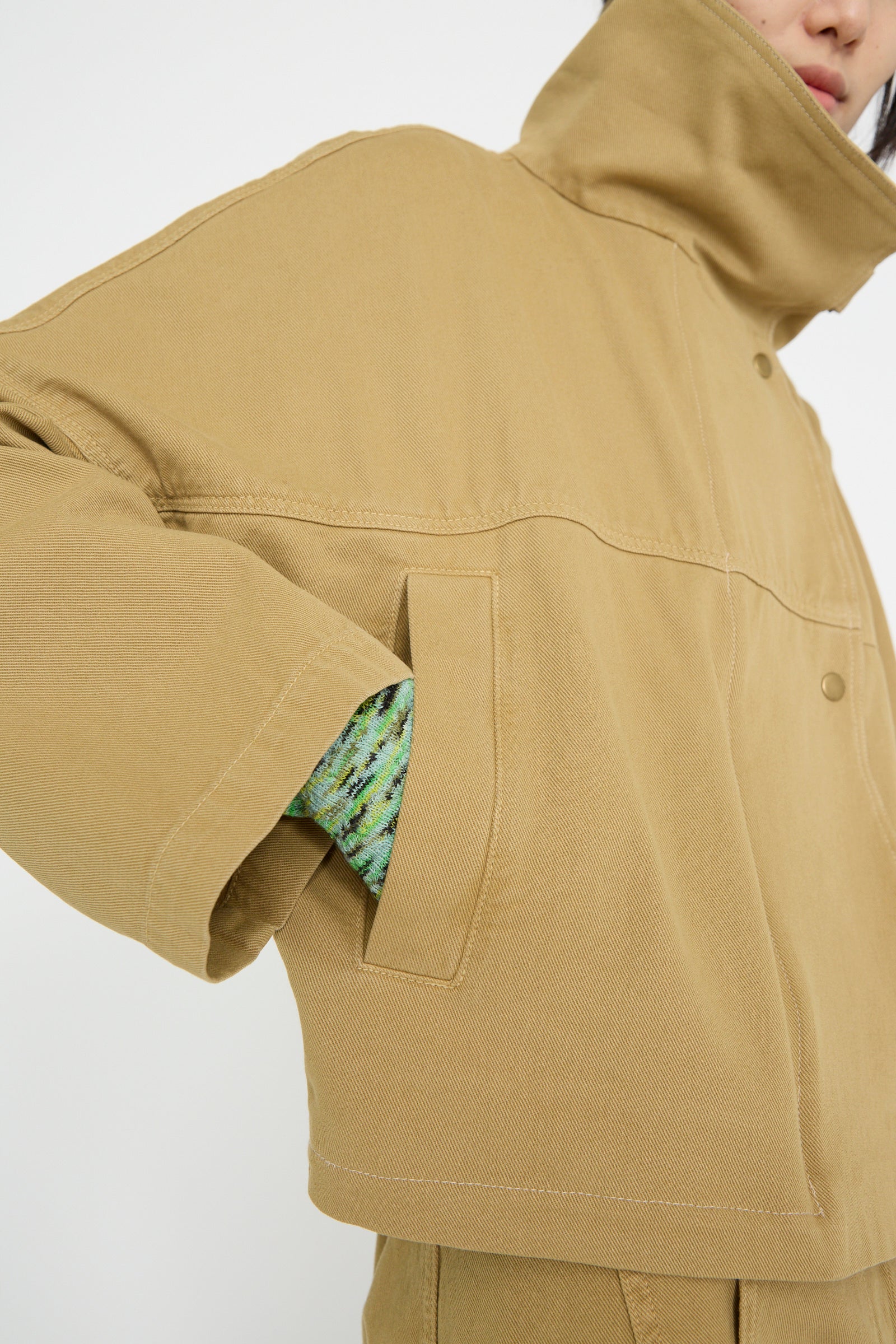 A person in a Vansire Jacket in Olive by Rachel Comey, cropped with a high collar and boxy fit, half-body view, wearing one hand in a pocket over a green patterned garment.