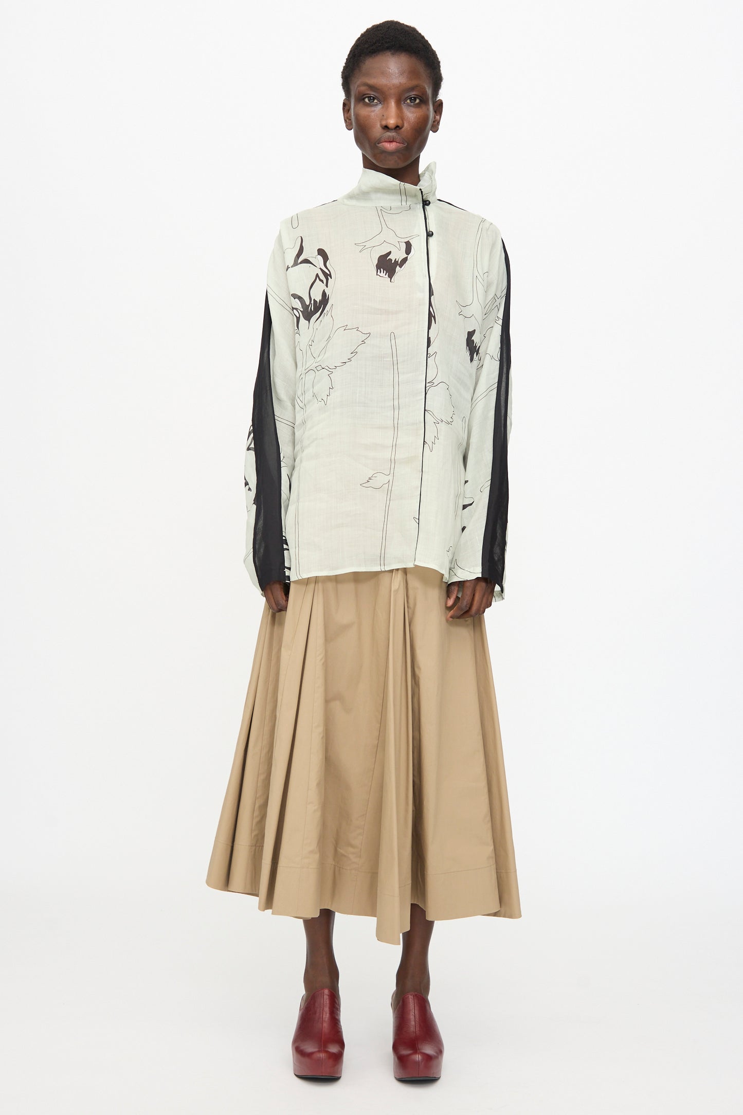 A person stands facing forward in the Rachel Comey Wanson Top in Dove, featuring a light floral print with black accents, paired with a beige pleated midi skirt and red heeled shoes against a plain white background.