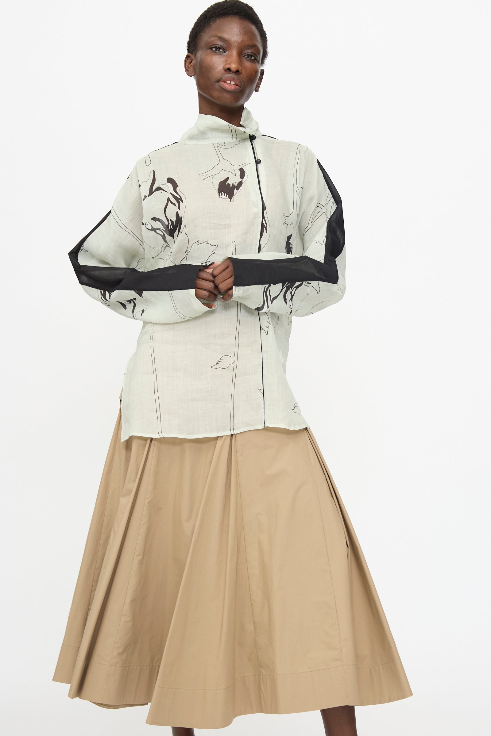 A person wears the Rachel Comey Wanson Top in Dove—a light-colored, long sleeve floral print shirt with black details—paired with a beige pleated midi skirt, standing against a plain white background.