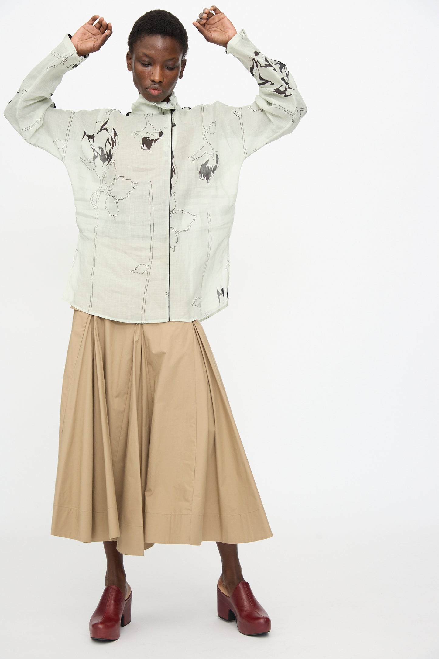 A person wearing the Rachel Comey Wanson Top in Dove, a beige pleated midi skirt, and red heeled shoes stands with arms raised against a plain white background.