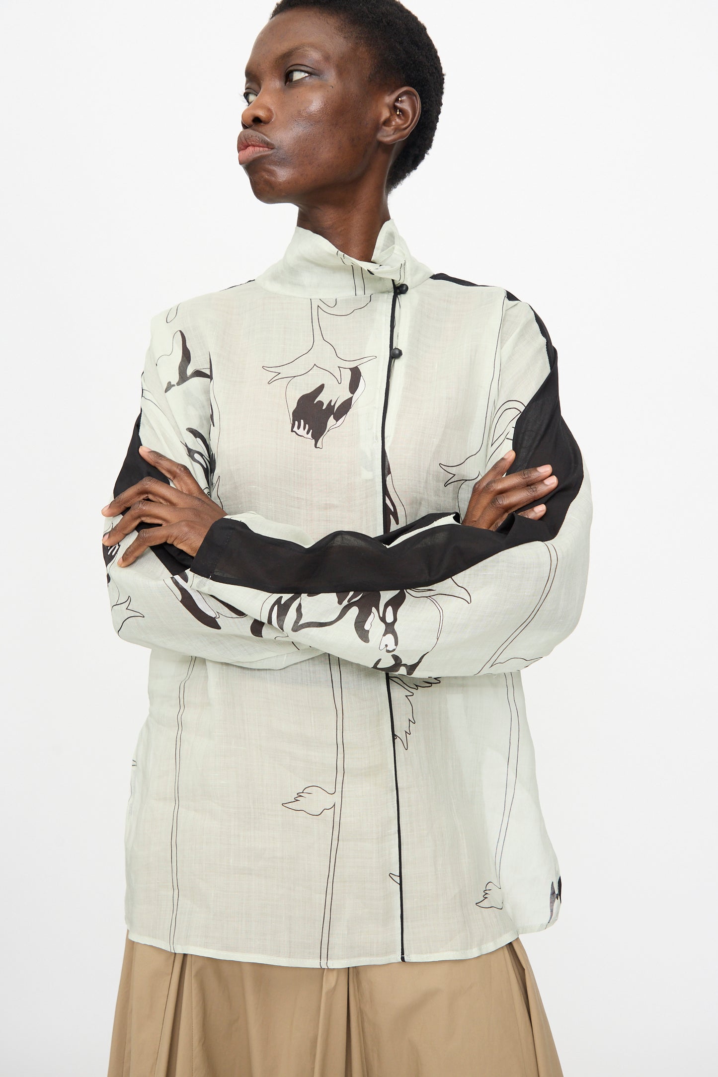 A person stands sideways with arms crossed, wearing the Rachel Comey Wanson Top in Dove—featuring abstract black line designs—and beige pants, set against a plain white background.