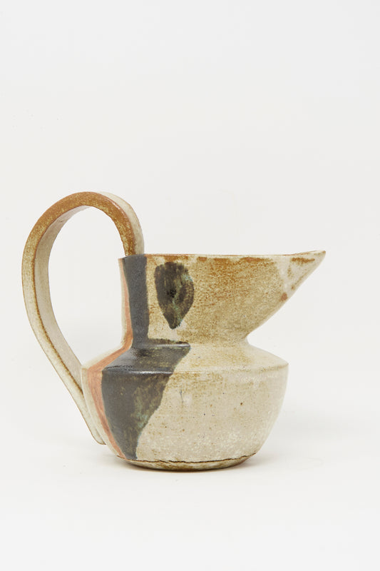 The Drawn Pitcher in Creme by Rachel Craven is handmade from wheel-thrown clay, featuring a wide handle and round body with abstract black and brown mineral glaze on a beige base, set against a plain white background.