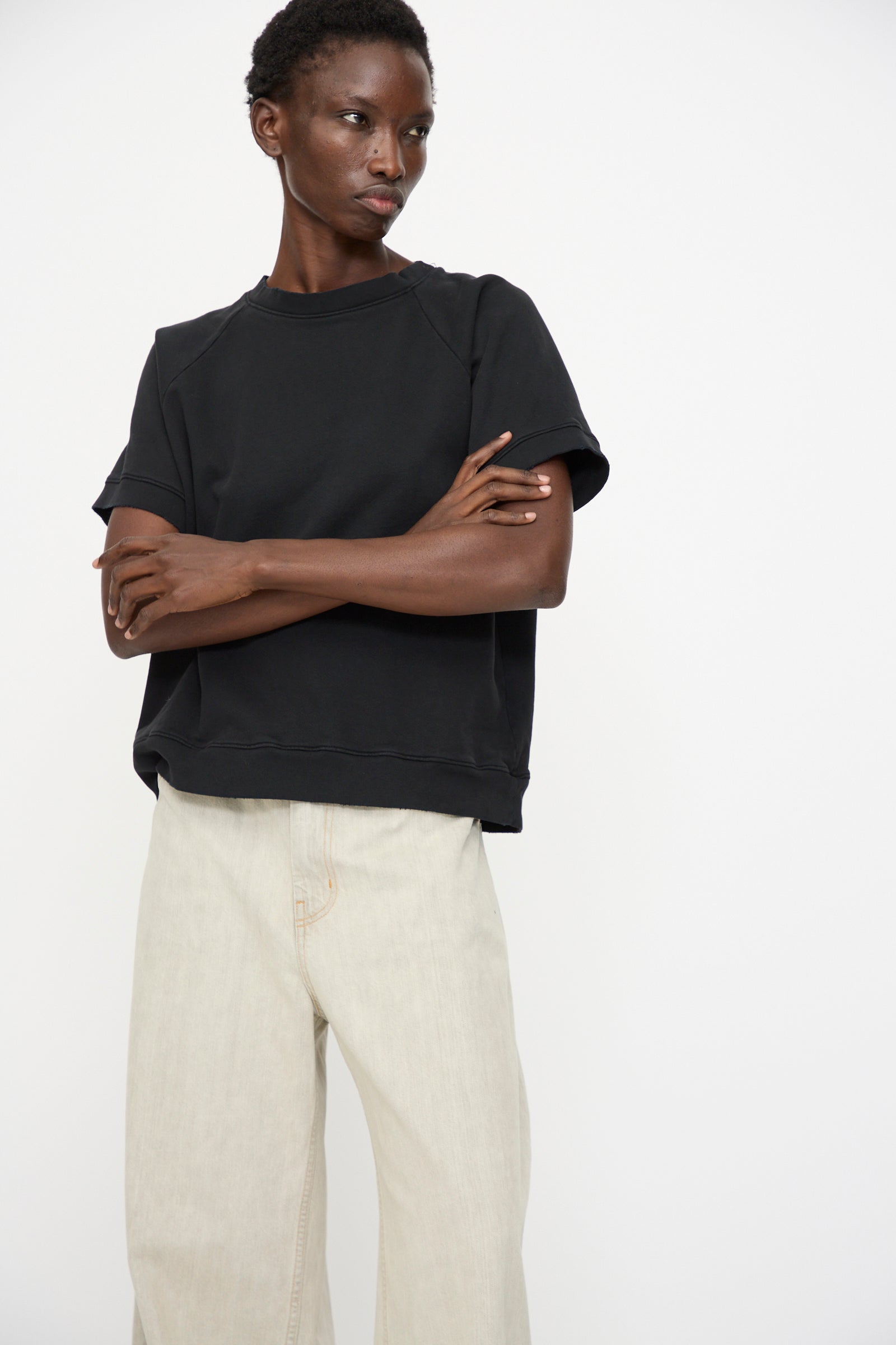 Person standing against a plain white background, arms crossed and neutral expression, wearing the Rachel Comey Billi Sweatshirt in Charcoal and light beige wide-leg pants.