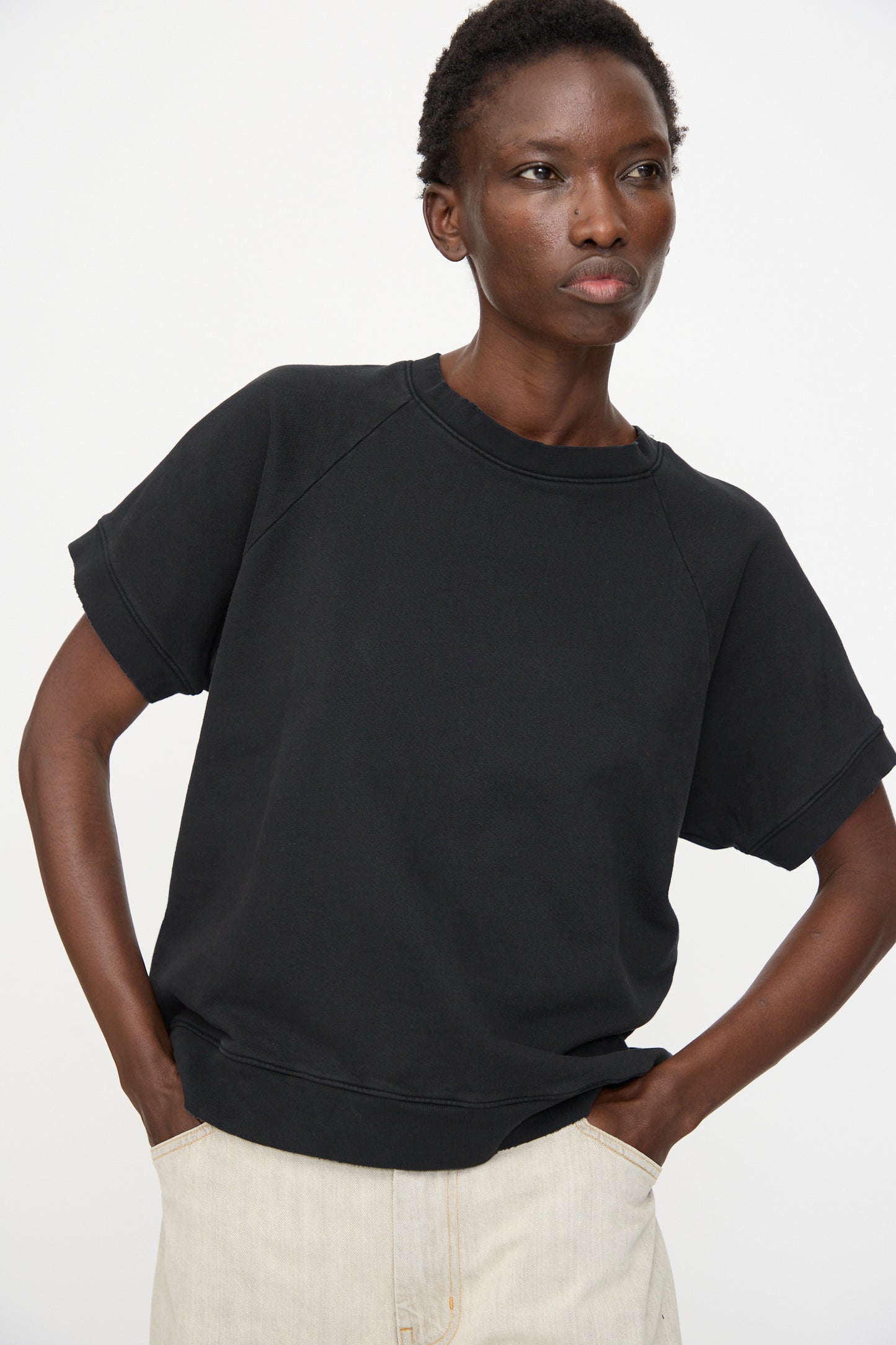 A person wears the Rachel Comey Billi Sweatshirt in Charcoal, a 100% cotton black crewneck short sleeve, paired with light-colored pants, standing with hands in pockets against a plain white background.