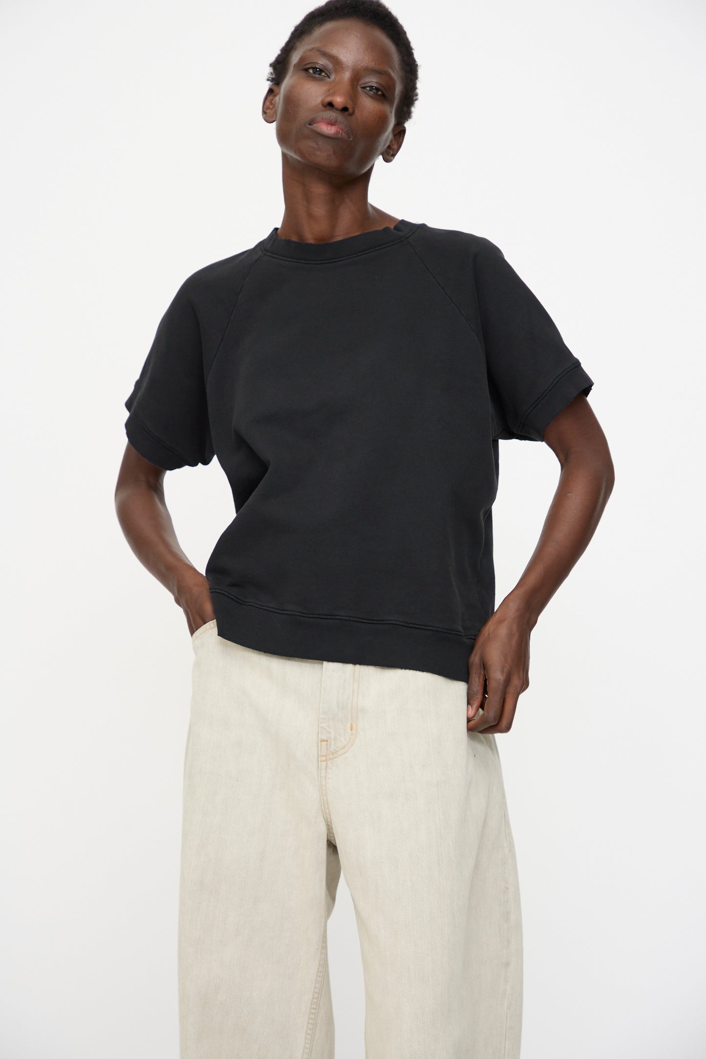 A person wears the Rachel Comey Billi Sweatshirt in Charcoal, a loose 100% cotton crewneck short sleeve top, paired with light beige wide-leg pants, standing against a plain white background.