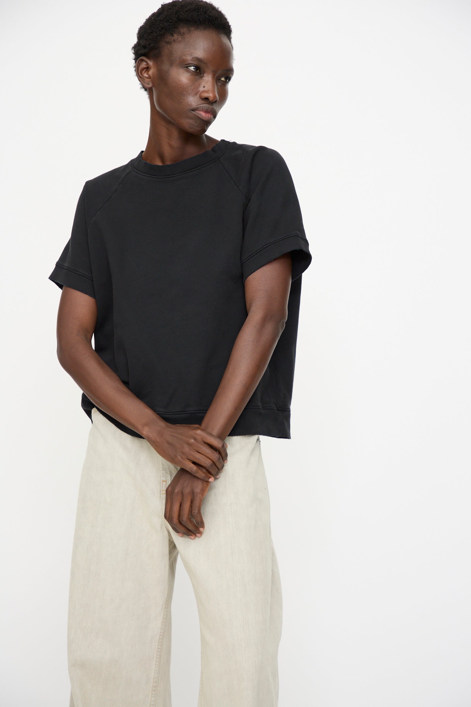 A person stands against a plain background, wearing the Rachel Comey Billi Sweatshirt in Charcoal (100% cotton crewneck, short sleeve) and light-colored wide-leg pants, hands clasped in front.