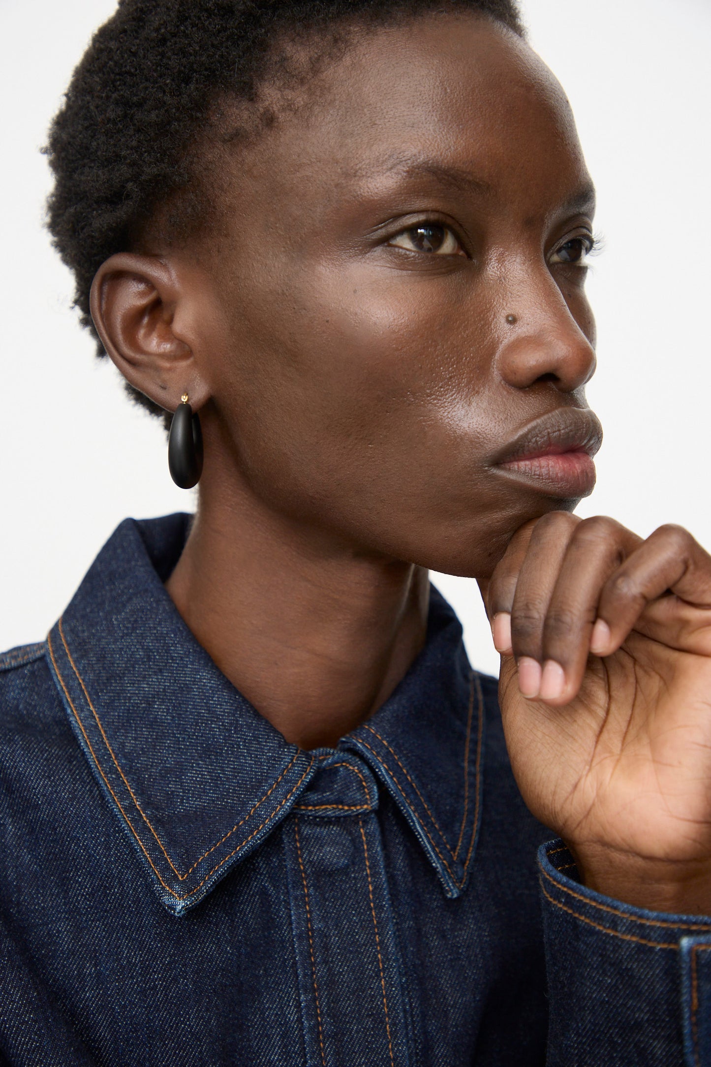 A person with short hair and a dark denim jacket poses thoughtfully with their hand on their chin, wearing Rachel Comey Grass Earrings in Black Matte against a plain background.