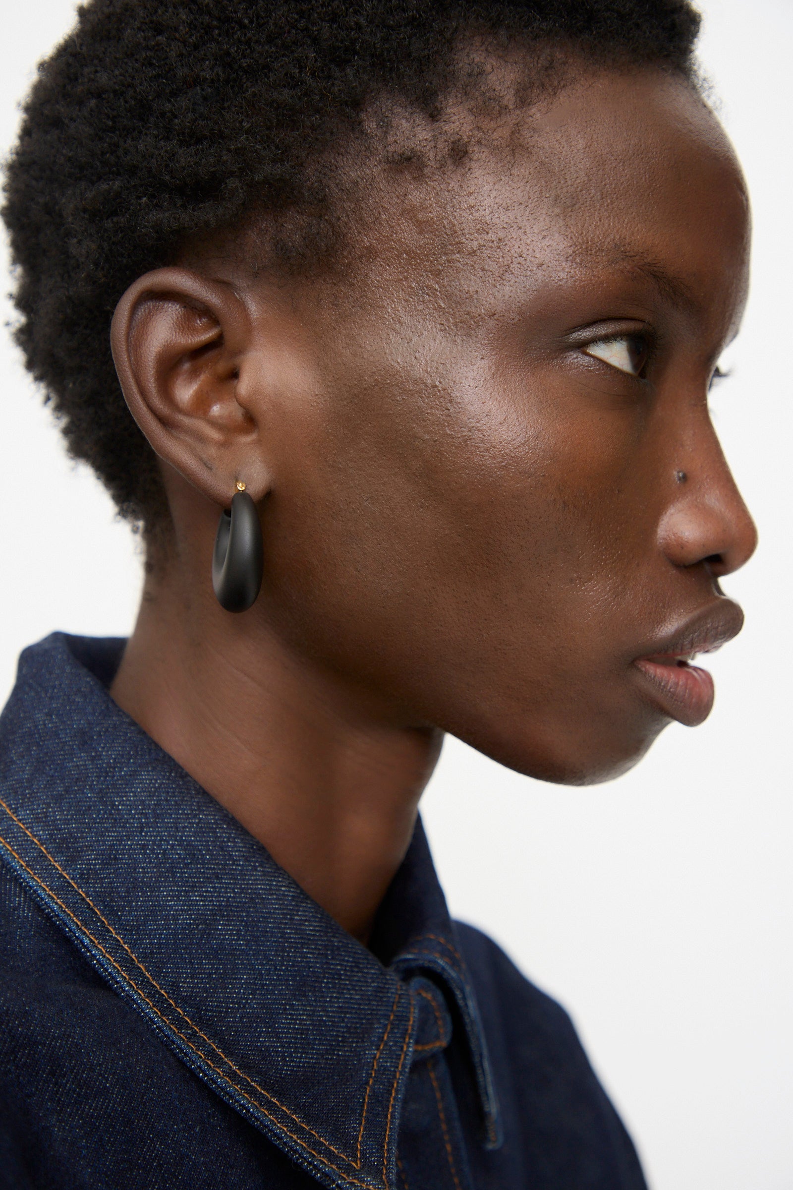 A person in a dark denim shirt, seen in right profile, wears Rachel Comey Grass Earrings in Black Matte and a nose stud against a plain background.