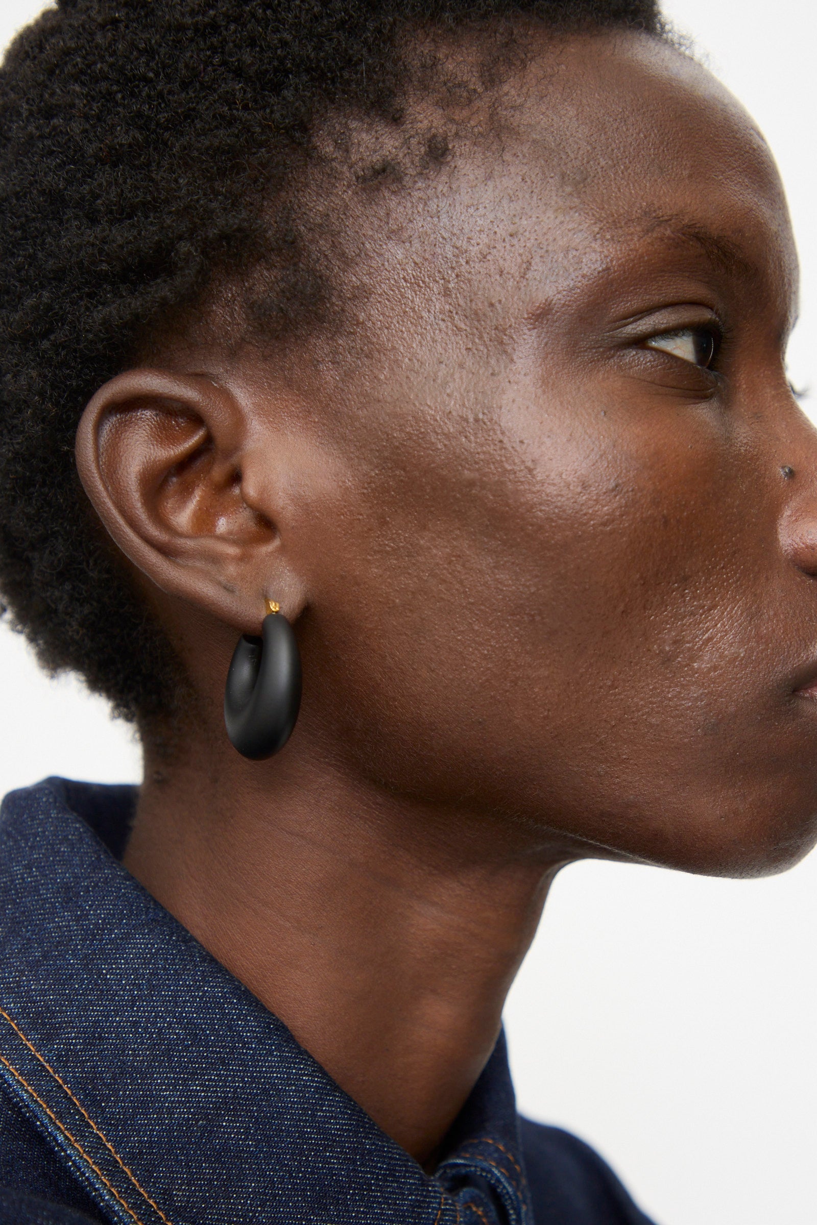 Close-up side view of a person with short hair wearing a blue denim shirt and Rachel Comey Grass Earrings in Black Matte.
