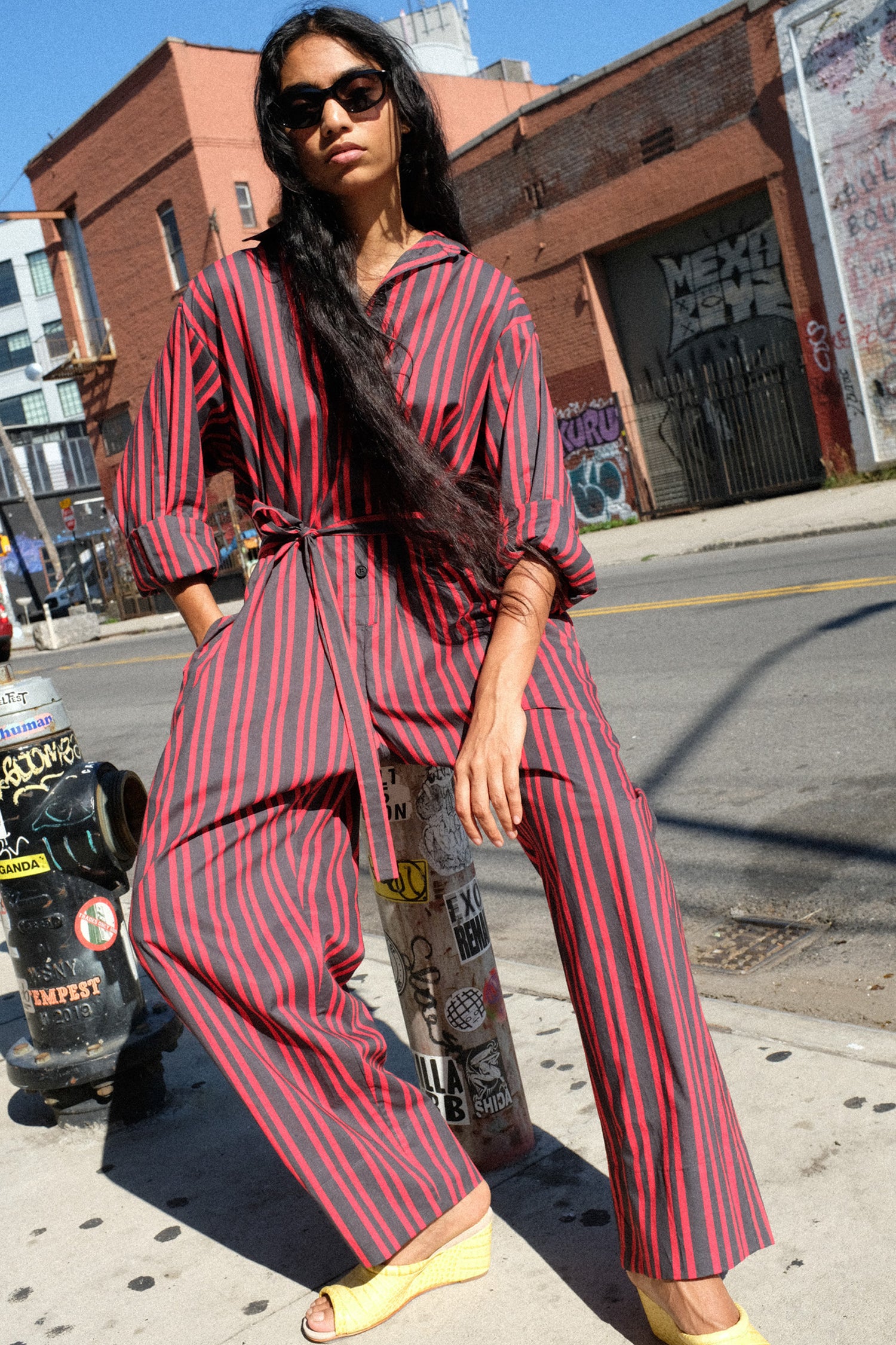 A woman wearing a red and black vertically striped jumpsuit with a tie waist, styled with yellow wedge sandals and sunglasses, posing on an urban city street.