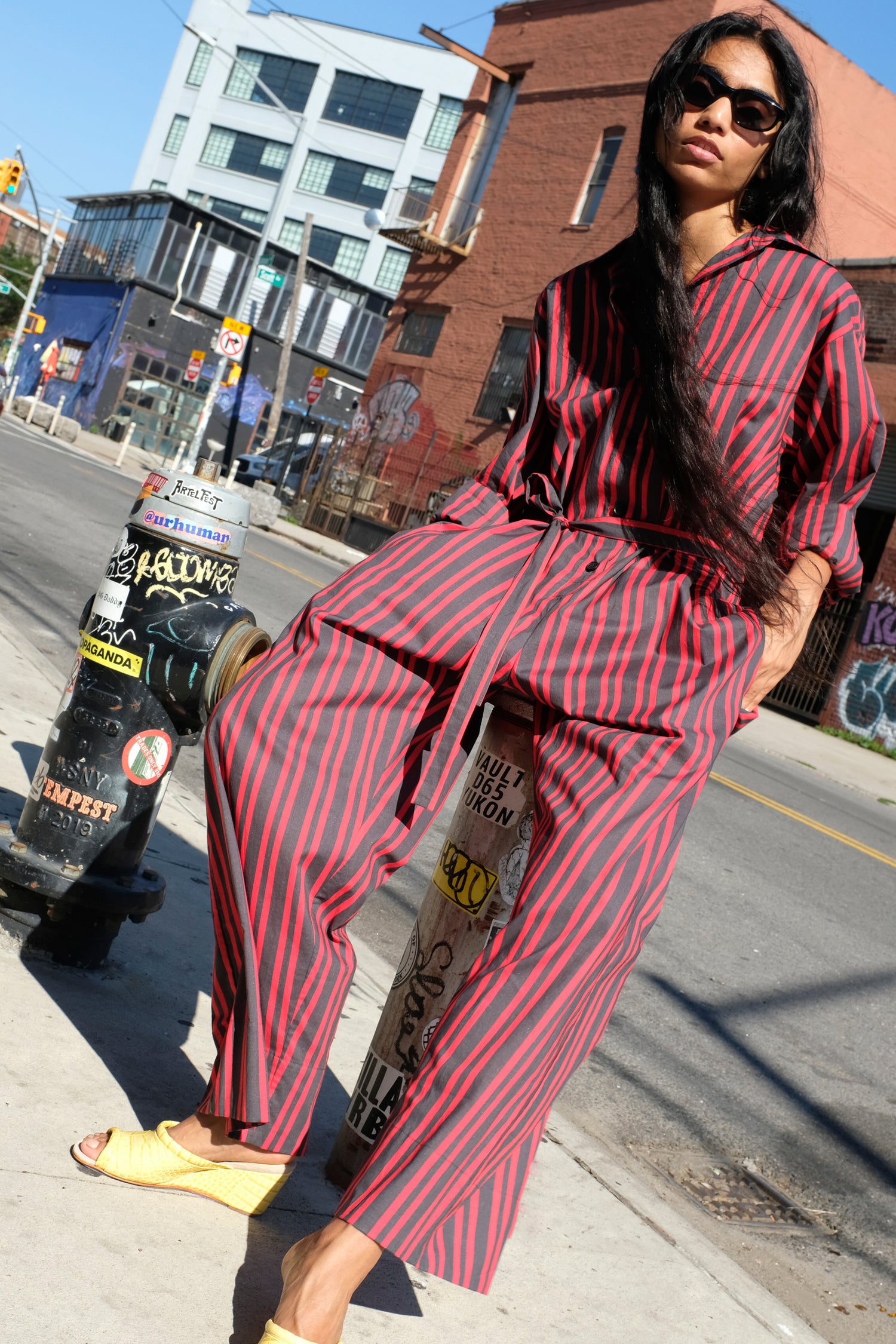 A woman wearing Rachel Comey's Smith Stripe Olli Jumpsuit in Red and Bully Mule in Banana poses on a city sidewalk next to a fire hydrant.