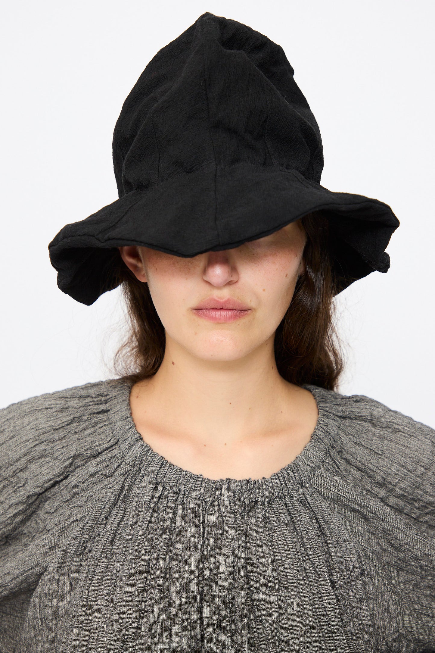 Wearing a textured gray top and the Reinhard Plank Cotton Ces Hat in Black, this individual stands against a plain white background, with the hat’s large brim covering their eyes.