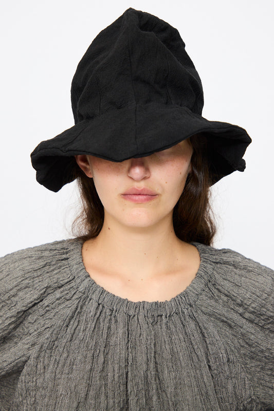 Wearing a textured gray top and the Reinhard Plank Cotton Ces Hat in Black, this individual stands against a plain white background, with the hat’s large brim covering their eyes.