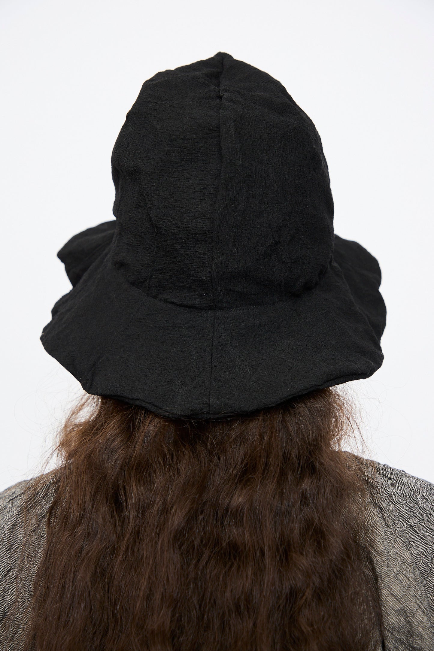 A person with long, wavy brown hair wears the Reinhard Plank Cotton Ces Hat in Black and a gray Italian-made top, photographed from behind against a plain background.