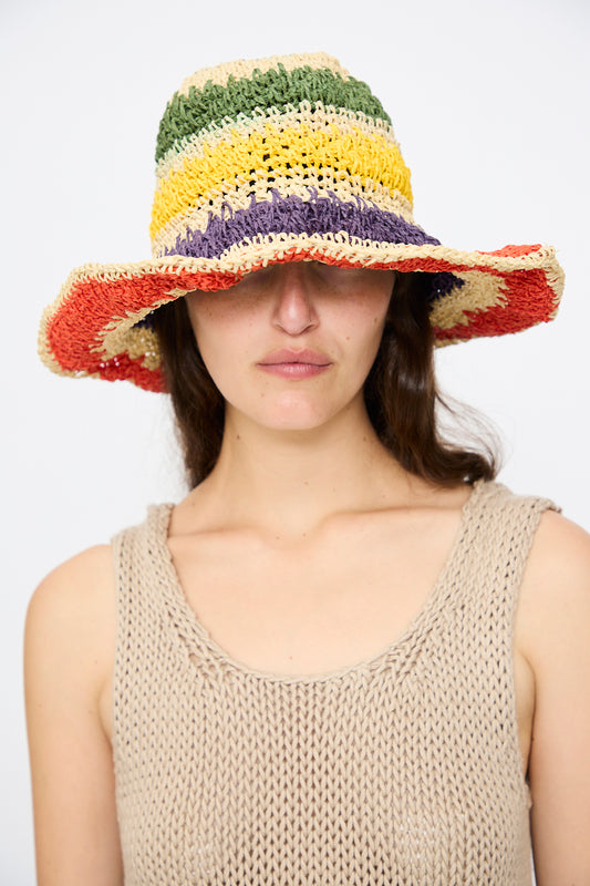 A woman wears a beige knit top and the Reinhard Plank Paper Unci Isle Hat in Stripes, a stylish foldable brim hat with multicolored straw that covers her eyes, standing against a plain background.