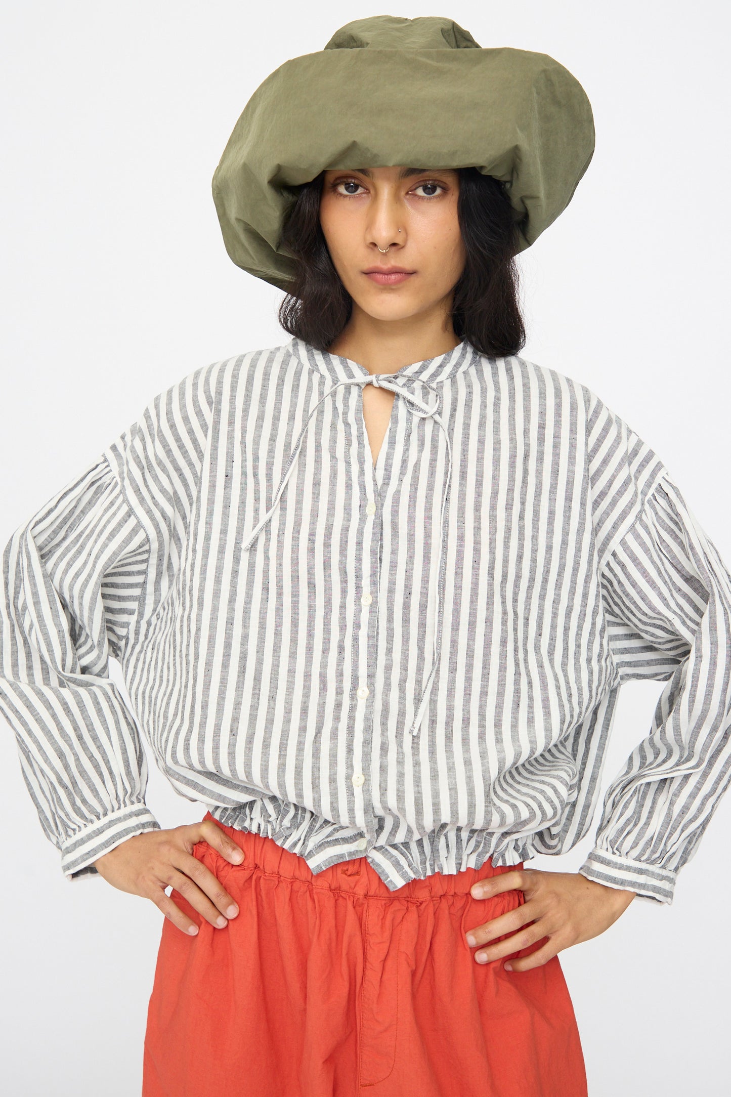 A person stands with hands on hips against a plain backdrop, wearing the Bianca Hat in Green by Reinhard Plank, a loose striped cotton button-up shirt with neck ties, and high-waisted red pants.