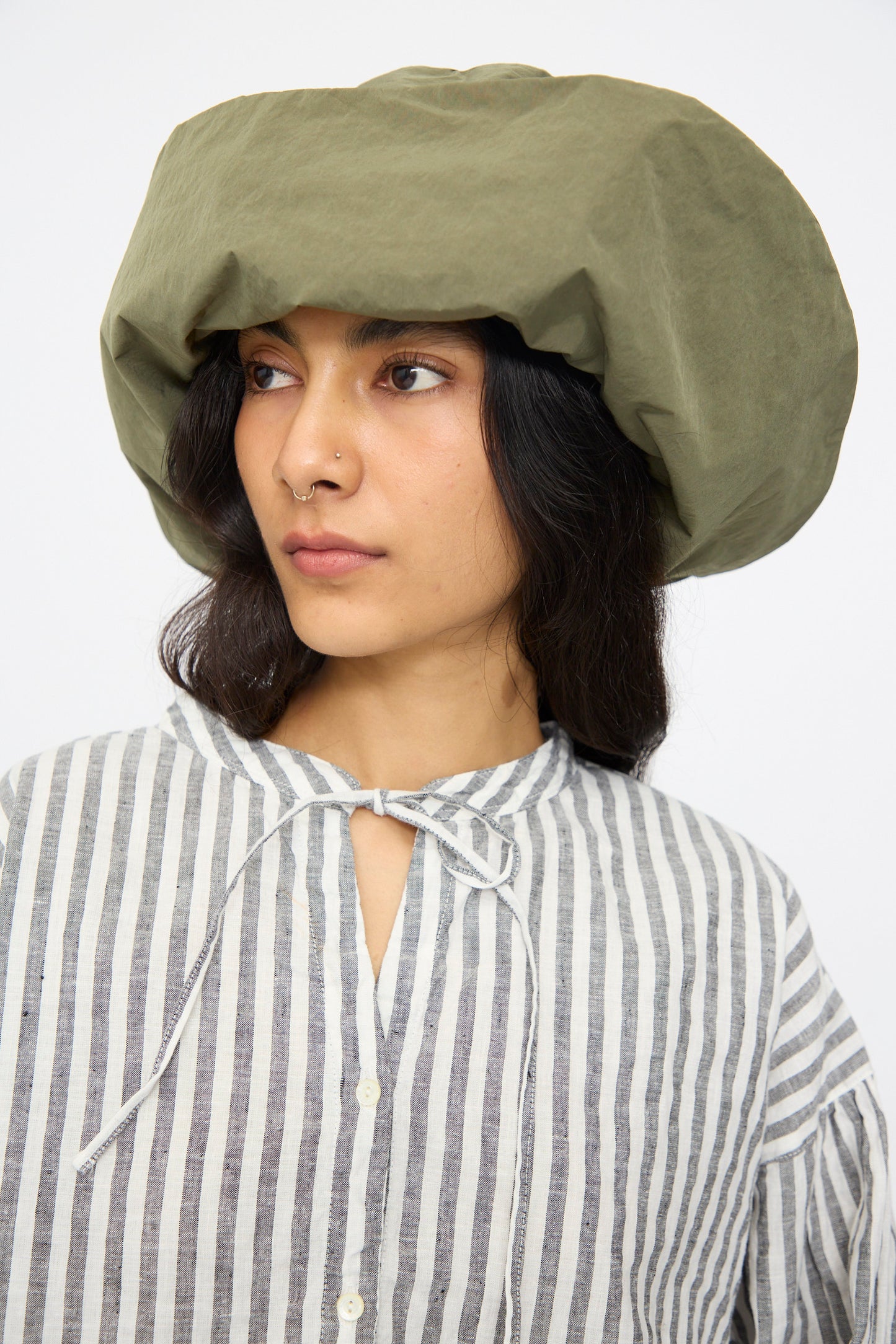 A person wearing the Bianca Hat in Green by Reinhard Plank and a striped, button-up shirt with a tied collar, looking slightly to the side.