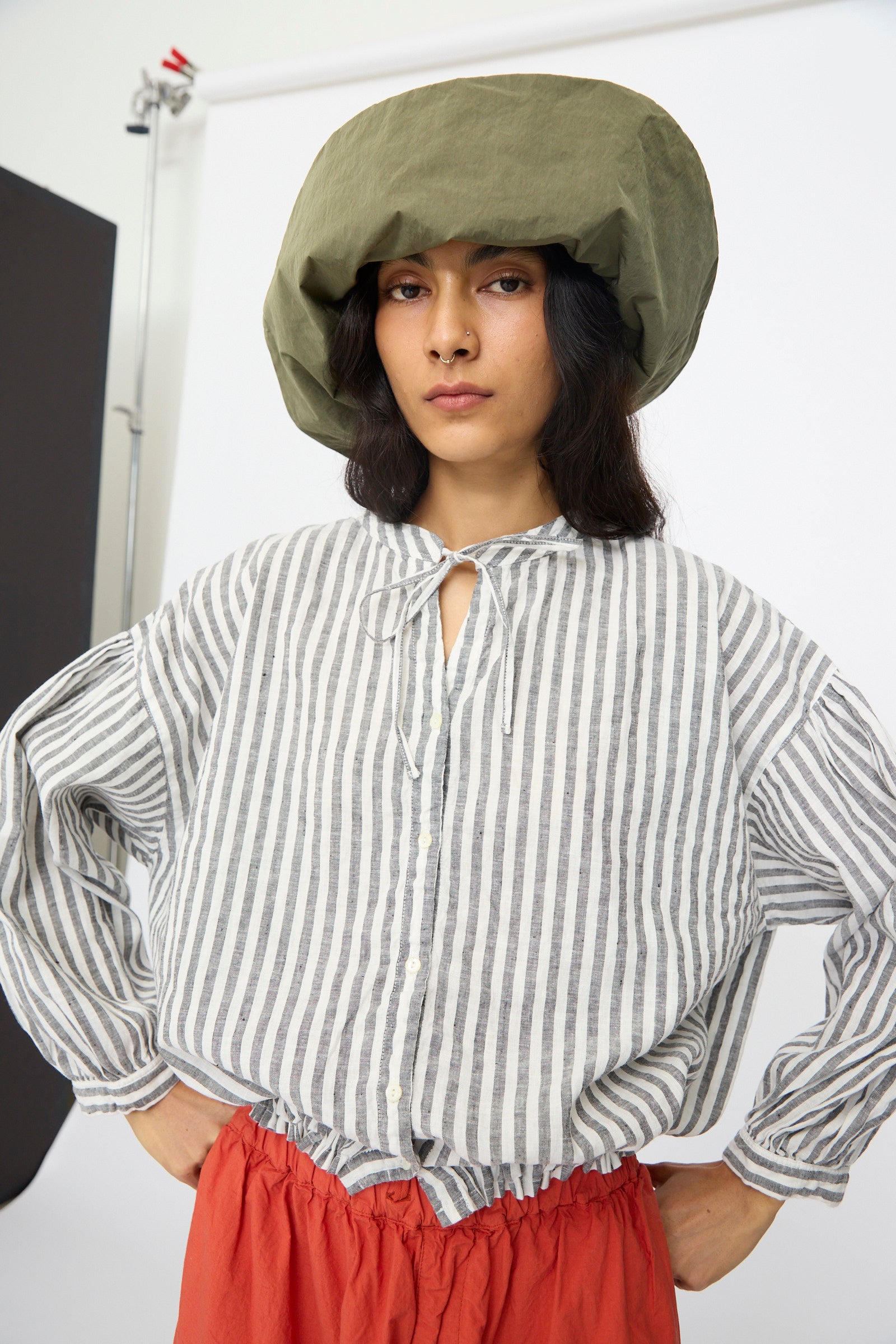 A person wears the Reinhard Plank Bianca Hat in Green, paired with a gray-and-white striped blouse and red pants, standing with hands on hips against a minimalist studio backdrop.