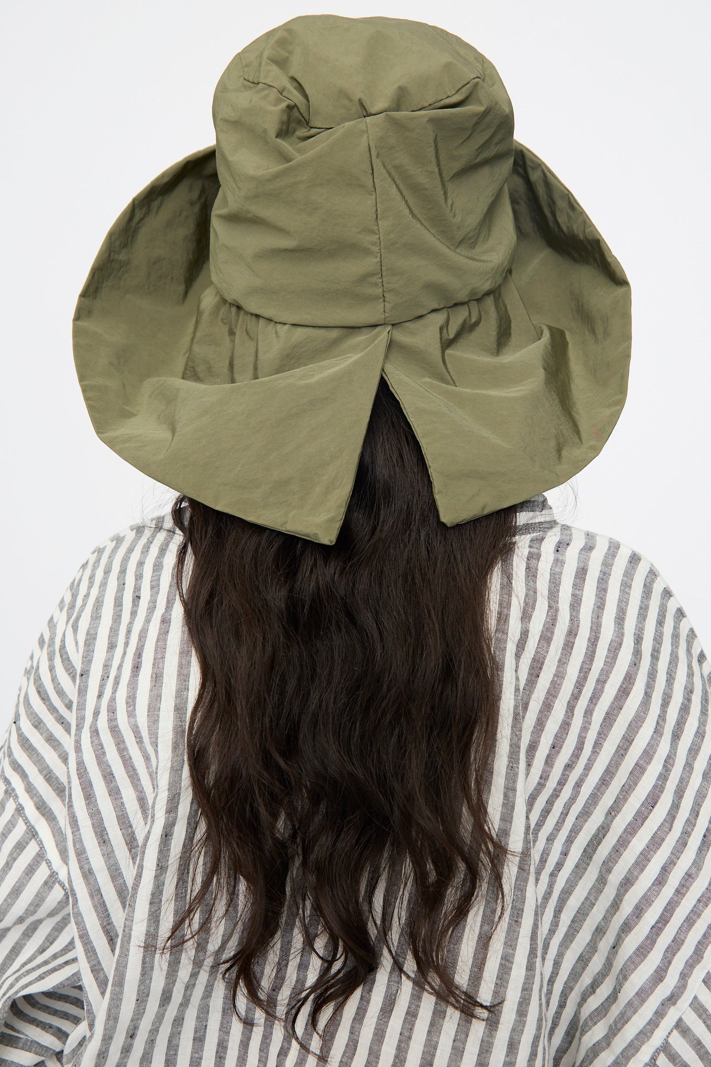 A person with long, wavy dark hair is seen from the back wearing the Reinhard Plank Bianca Hat in Green and a striped shirt against a plain background.