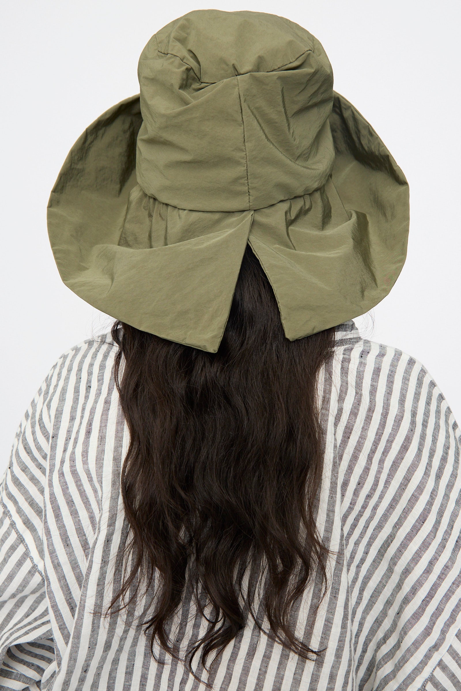 A person with long, wavy dark hair is seen from the back wearing the Reinhard Plank Bianca Hat in Green and a striped shirt against a plain background.