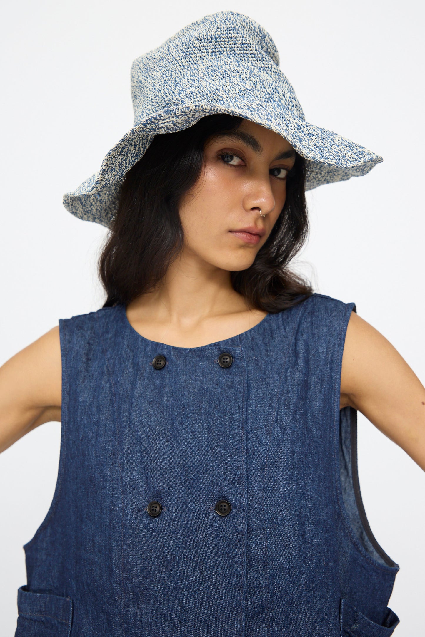 A woman wearing the Reinhard Plank Isle New Paper Hat in white and blue poses against a plain white background, paired with a sleeveless blue denim dress.