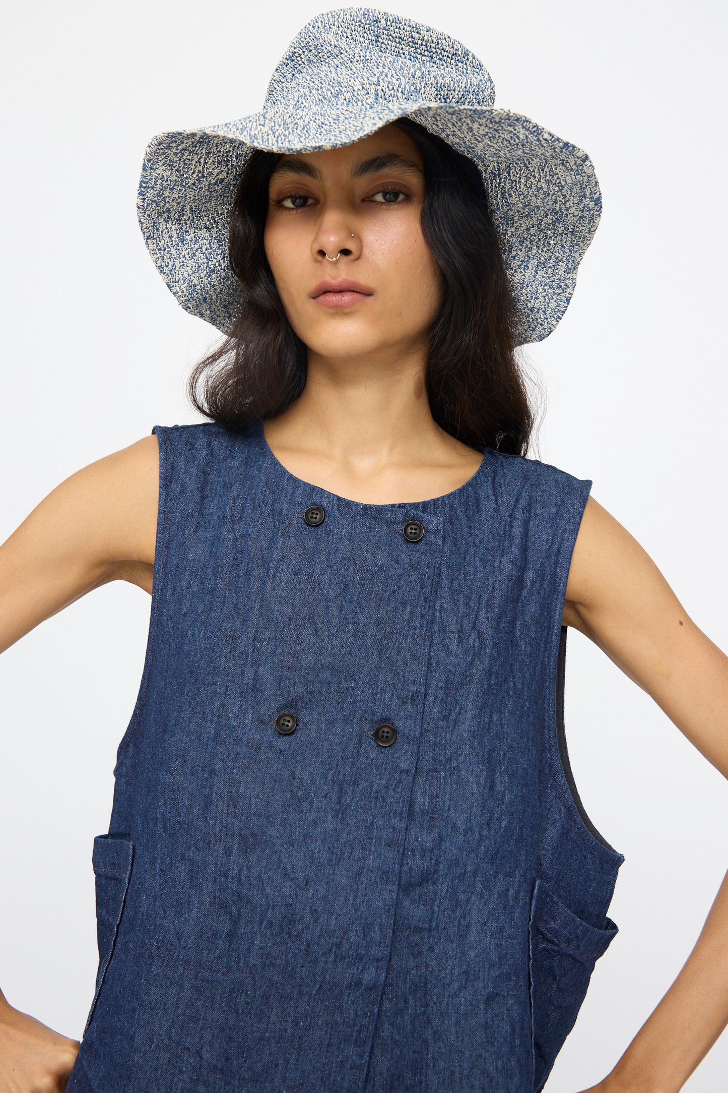 A woman wears the Reinhard Plank Isle New Paper Hat in White and Blue with a denim sleeveless dress featuring four black buttons.