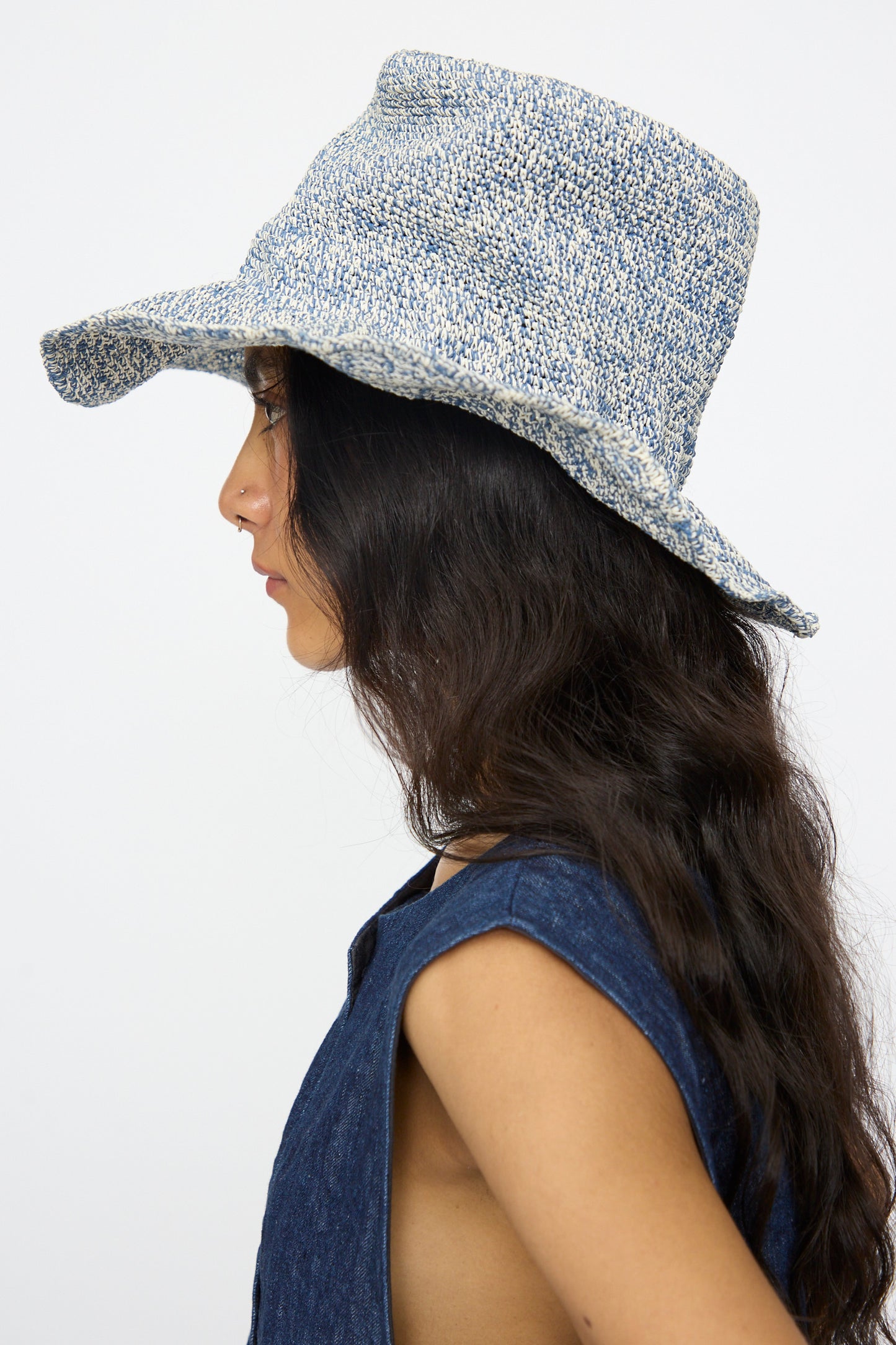A person with long dark hair, shown in profile against a plain background, wears the Isle New Paper Hat in White/Blue by Reinhard Plank and a sleeveless blue top.