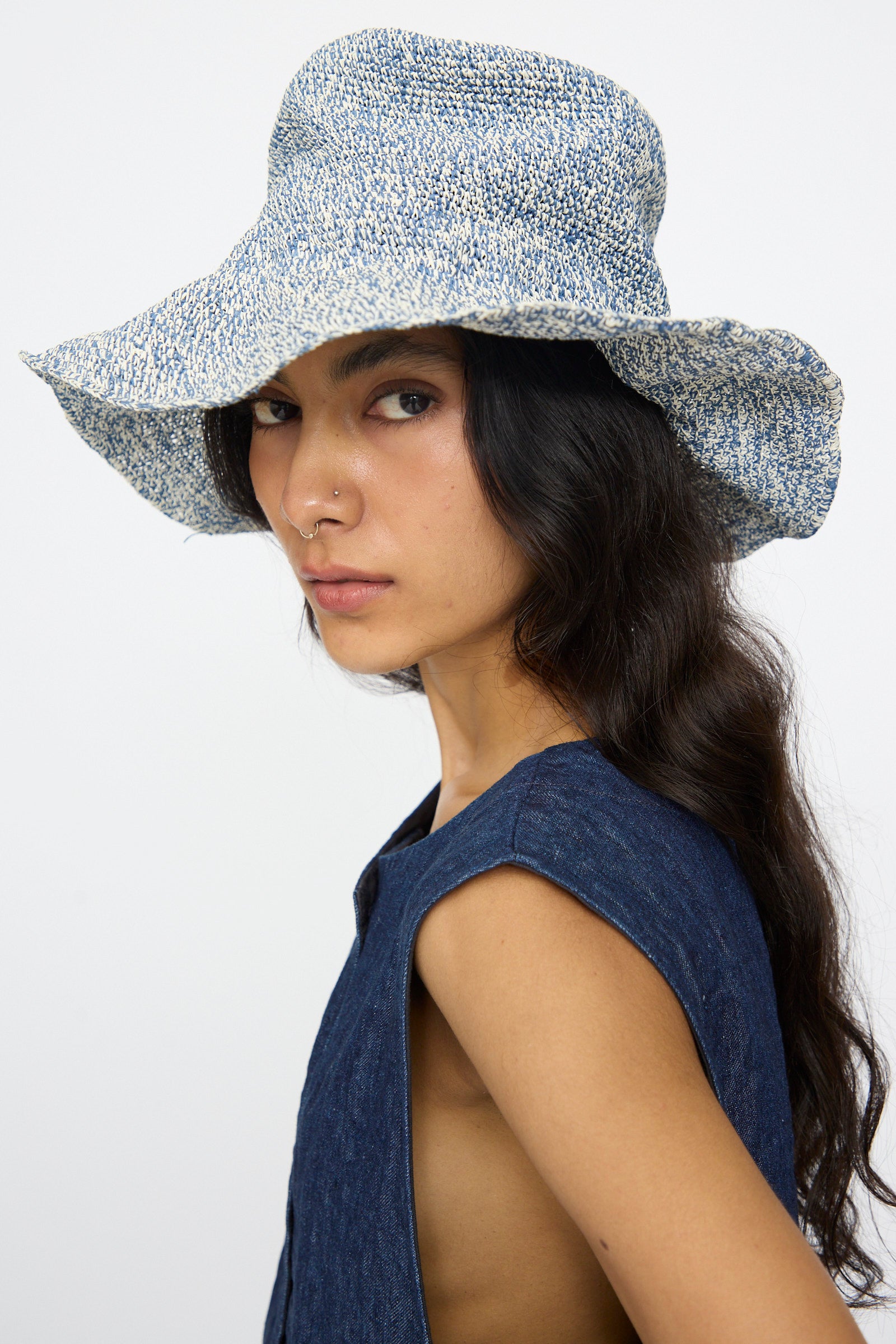 A person wearing the Isle New Paper Hat in White, Blue by Reinhard Plank and a denim vest, looks to the side.