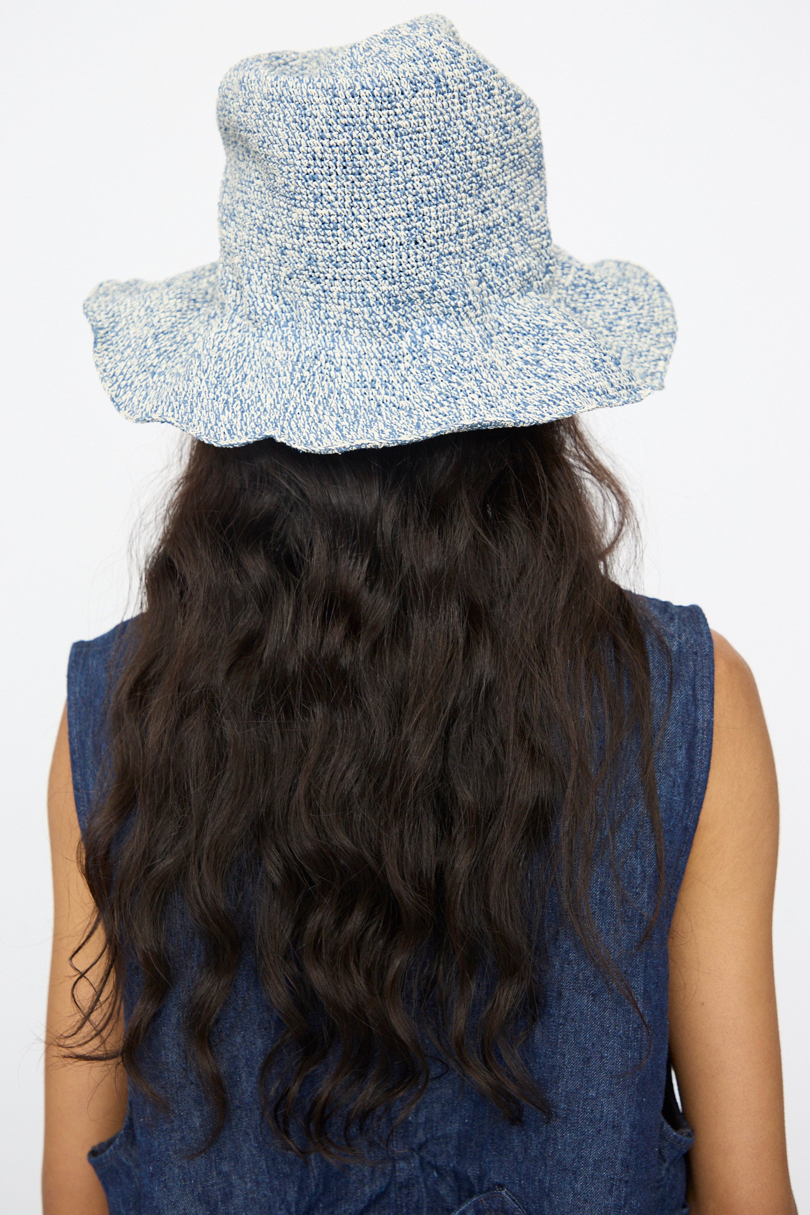 Person with long, wavy dark hair wears the Reinhard Plank Isle New Paper Hat in White, Blue and a sleeveless denim top, shown from the back against a plain white background.