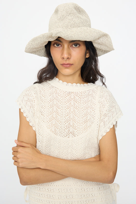 A woman wearing the Isle New Paper Hat in White by Reinhard Plank, featuring a wide-brimmed crocheted design, stands with arms crossed against a plain background, paired with a short-sleeve lace top.