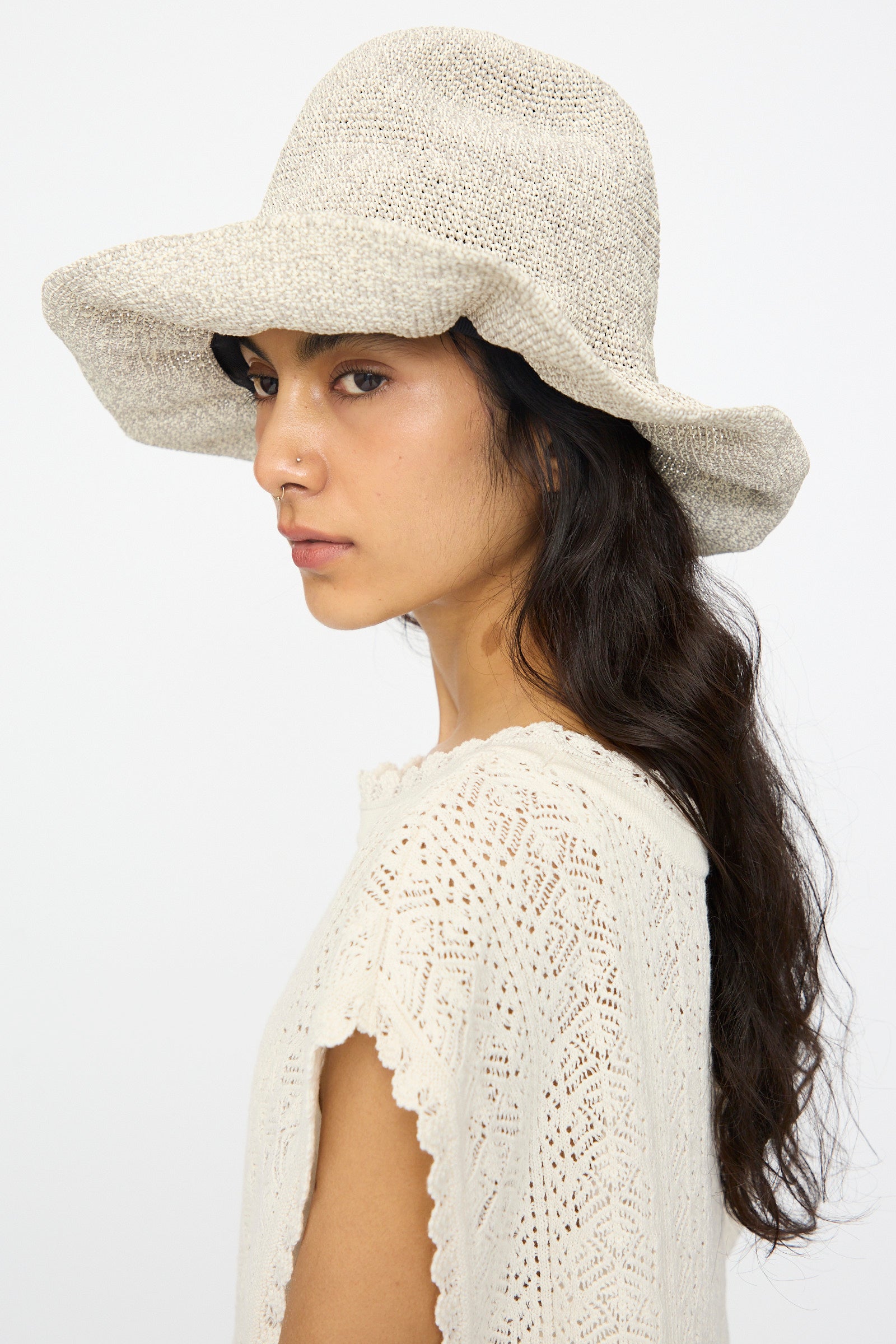 A woman with long dark hair in side profile wears the Isle New Paper Hat in White, Sand by Reinhard Plank and a cream crochet top, set against a plain background. 