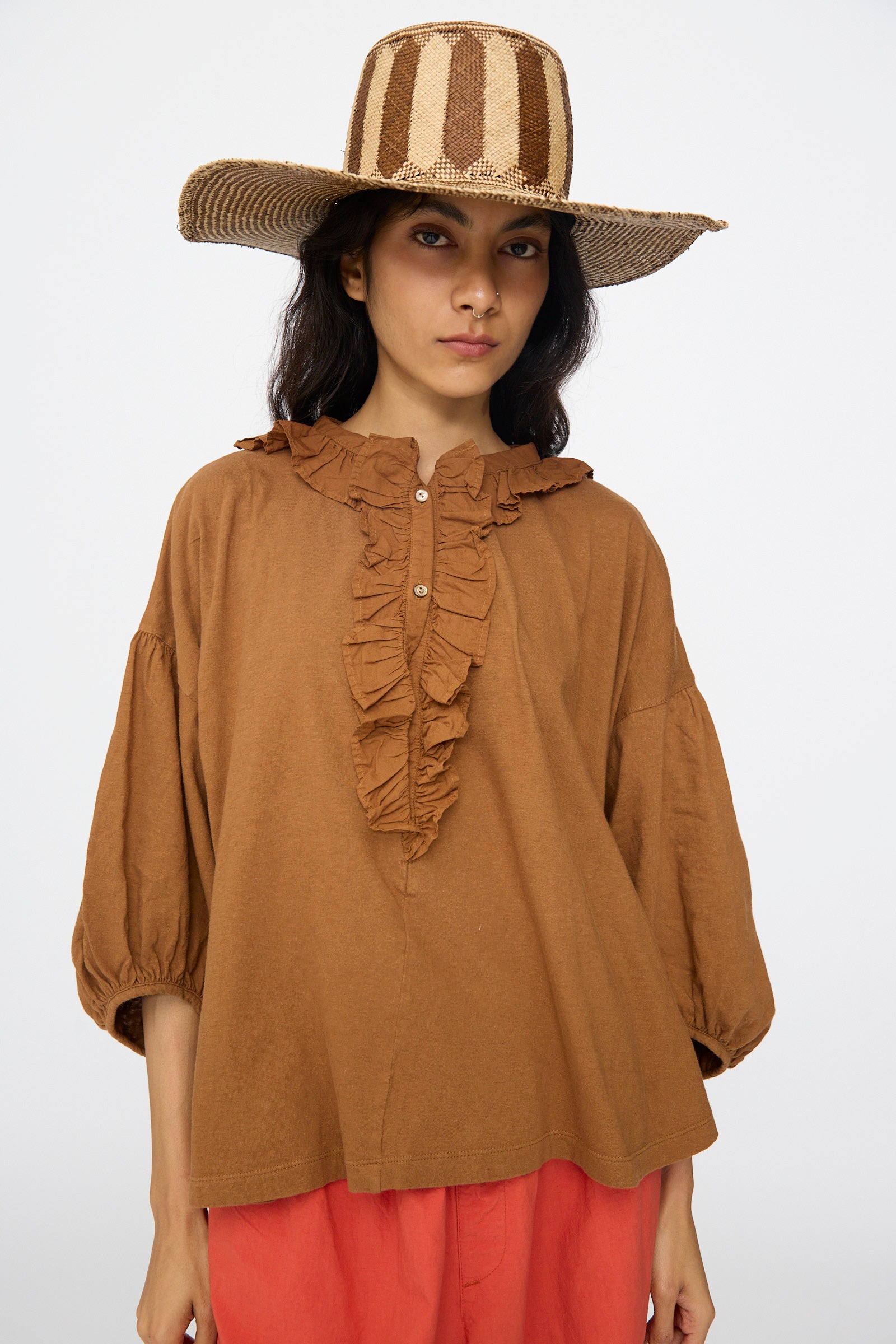A person wearing a wide-brim Straw Beghe Lai Hat in Natural Stripes by Reinhard Plank, a brown ruffled top, and coral-colored pants stands against a plain light background. 