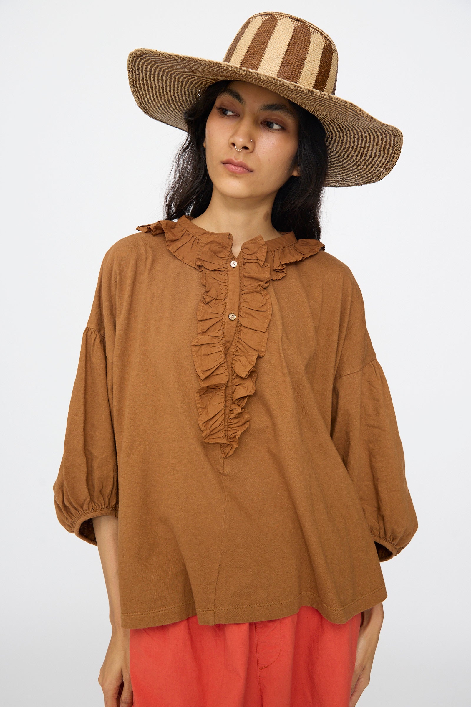 A woman wears the Reinhard Plank Straw Beghe Lai Hat in Natural Stripes, a brown ruffled button-up blouse, and coral pants while standing against a plain background.