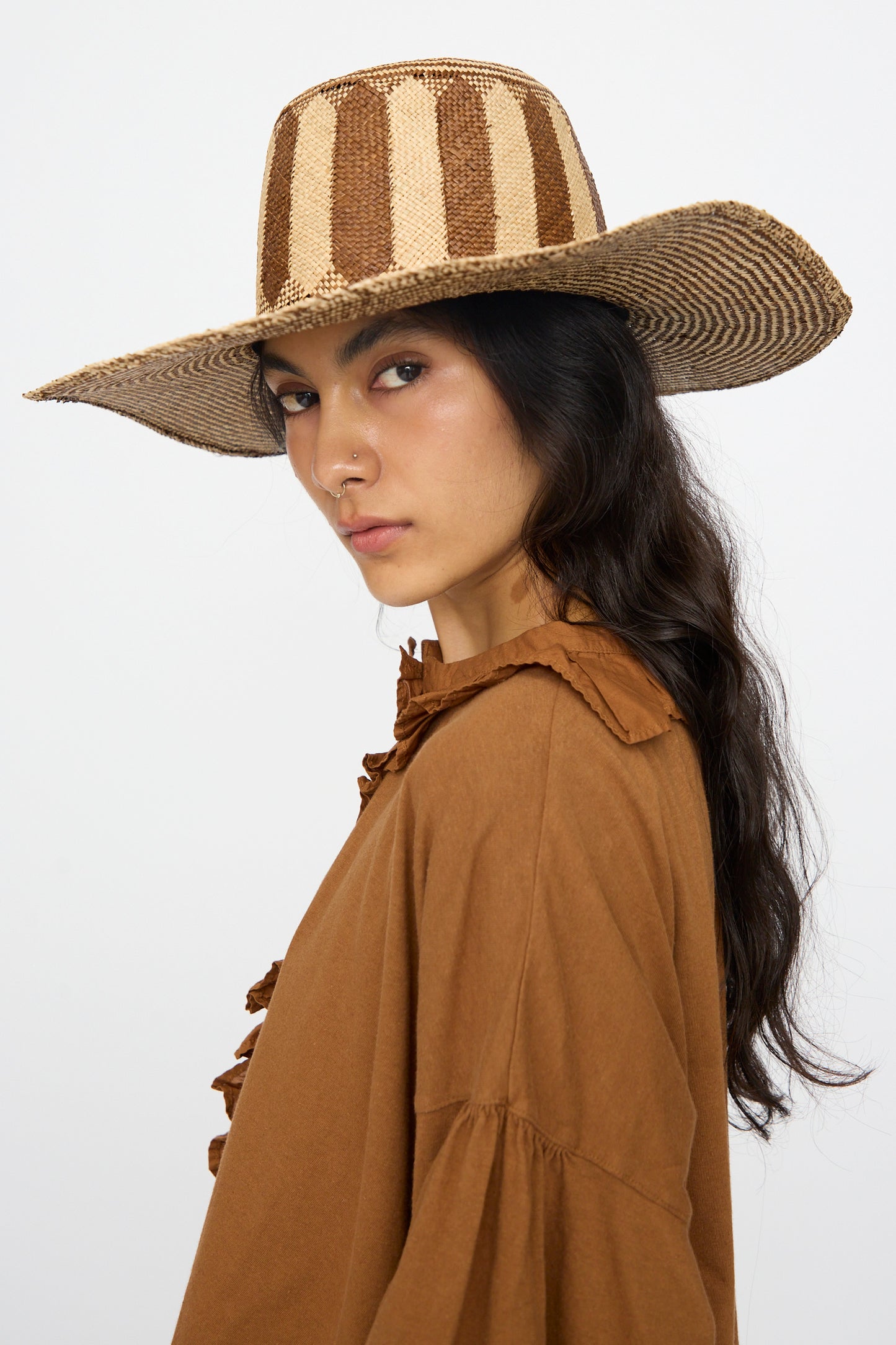 A woman wearing the Reinhard Plank Straw Beghe Lai Hat in Natural Stripes and a brown top stands against a white background.