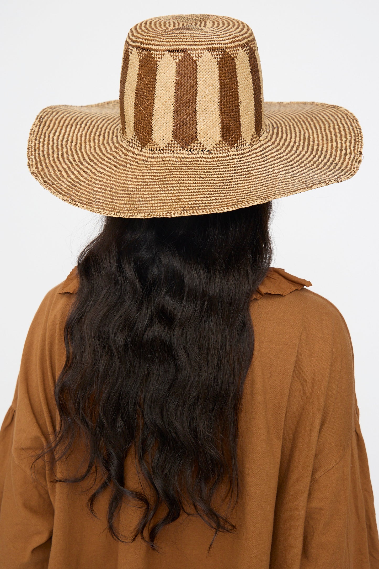 Person with long, wavy black hair wears the Reinhard Plank Straw Beghe Lai Hat in Natural Stripes and a brown shirt, seen from behind against a plain background.