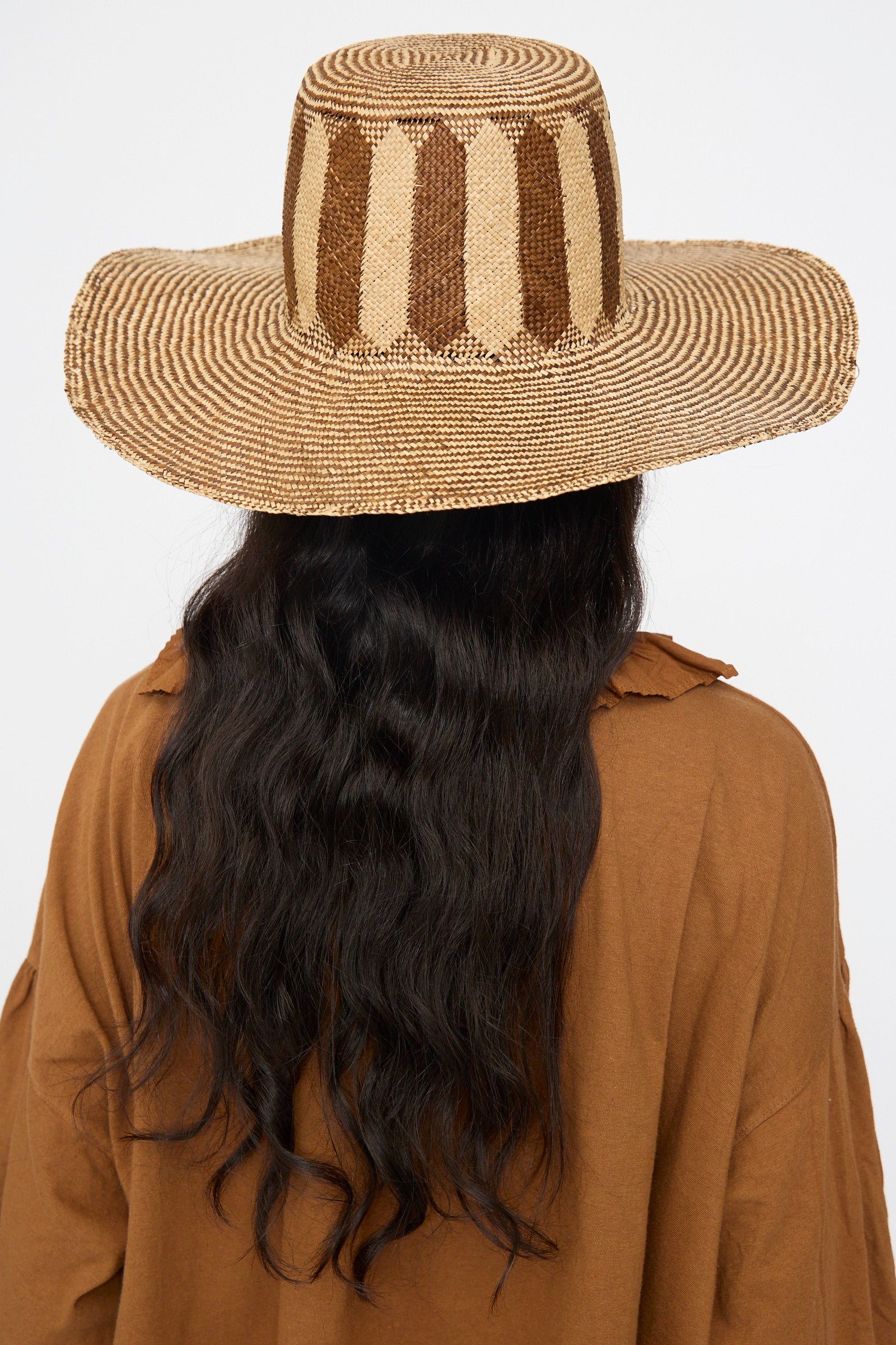 Person with long, wavy black hair wears the Reinhard Plank Straw Beghe Lai Hat in Natural Stripes and a brown shirt, seen from behind against a plain background.