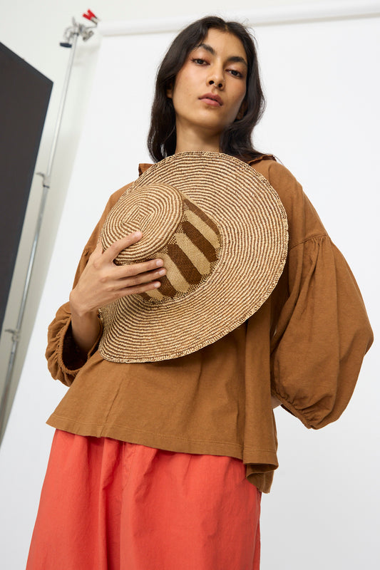 A woman holds the Reinhard Plank Straw Beghe Lai Hat in Natural Stripes against her chest, standing in front of a white backdrop while wearing a brown blouse and red pants.