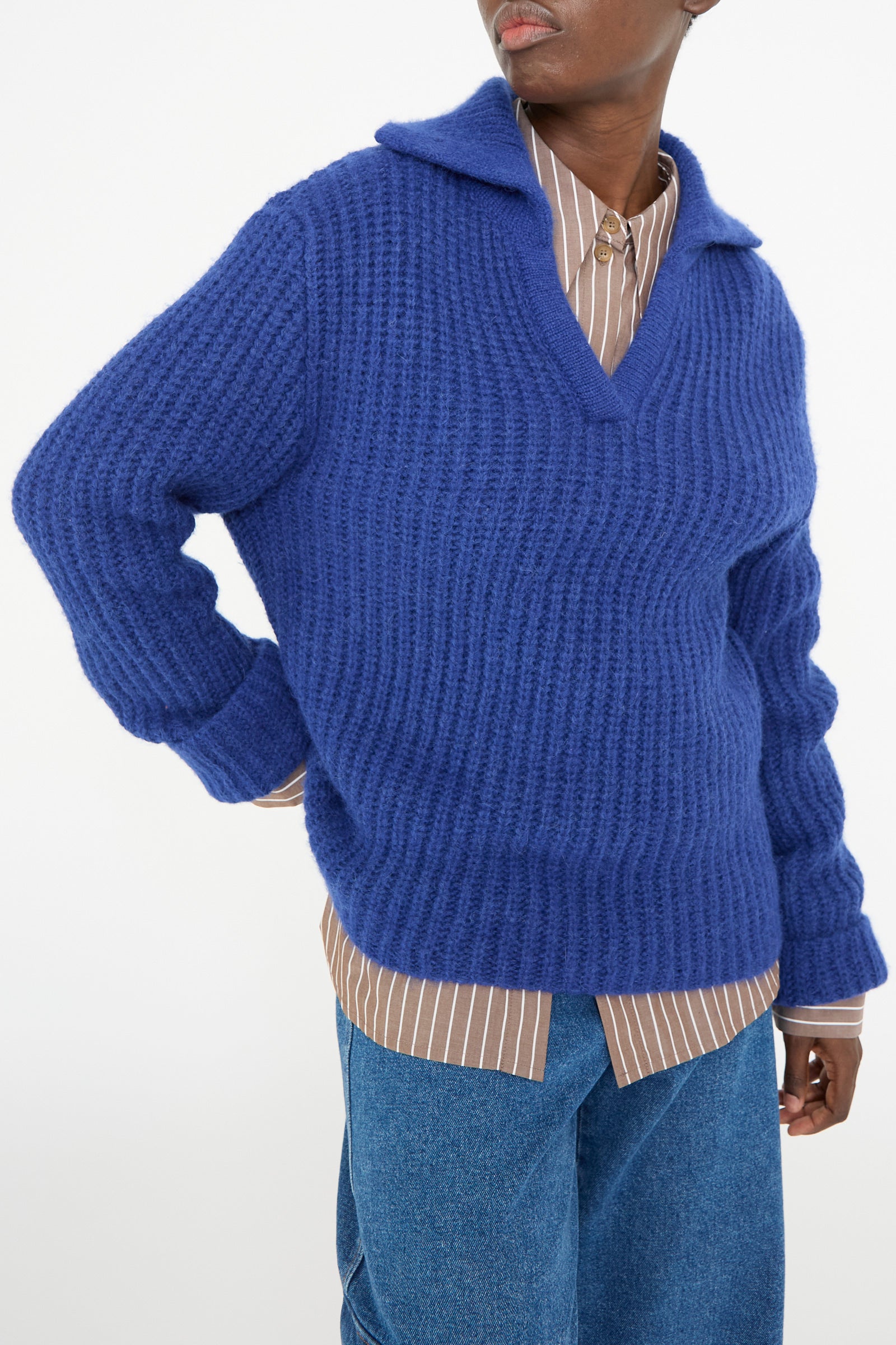 A person stands against a plain background, wearing the Rejina Pyo Chunky Alpaca Alison Sweater in Electric Blue over a brown striped shirt and blue jeans.