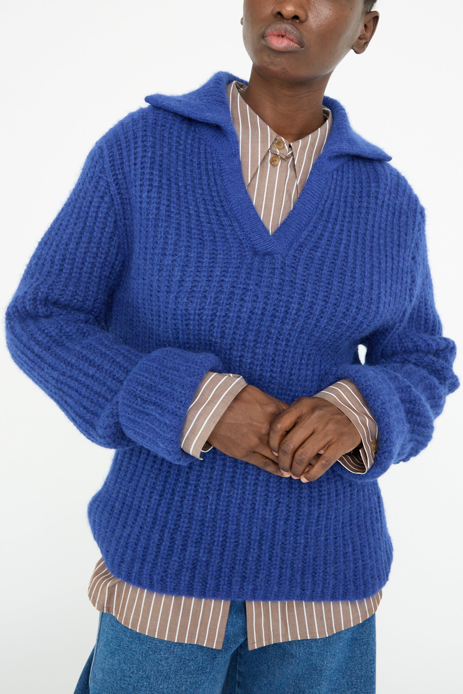 Wearing a Chunky Alpaca Alison Sweater in Electric Blue by Rejina Pyo over a brown striped shirt and blue jeans, a person stands against a white background.