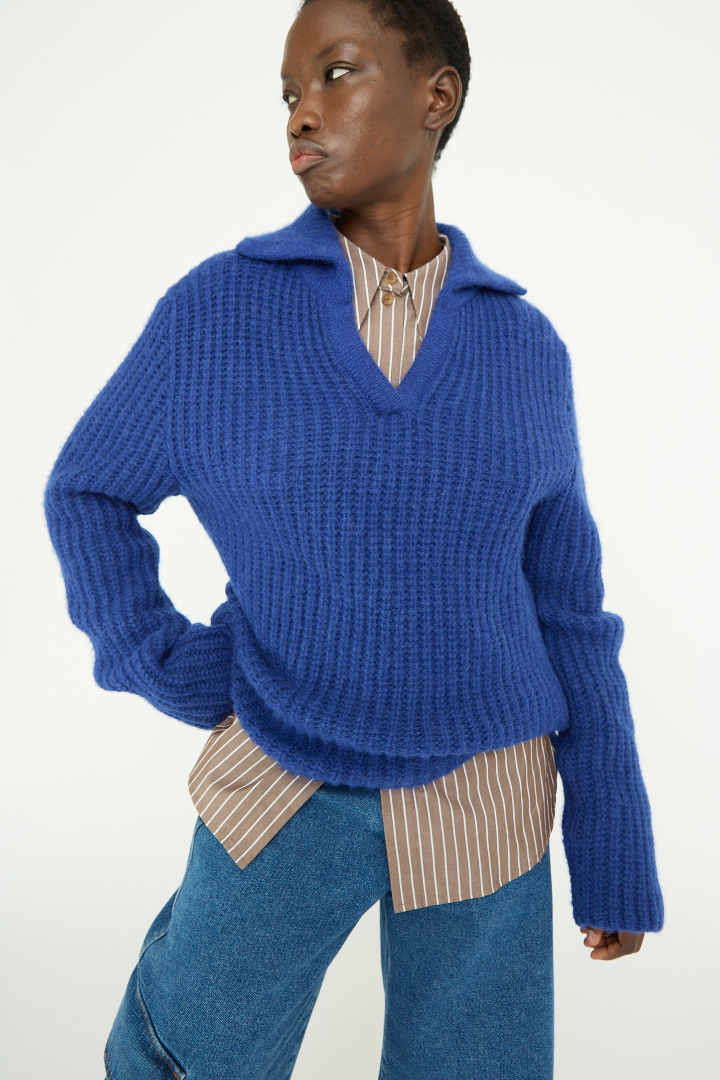 A person wearing a Rejina Pyo Chunky Alpaca Alison Sweater in electric blue over a brown striped shirt poses with one hand on their hip, looking to the left.