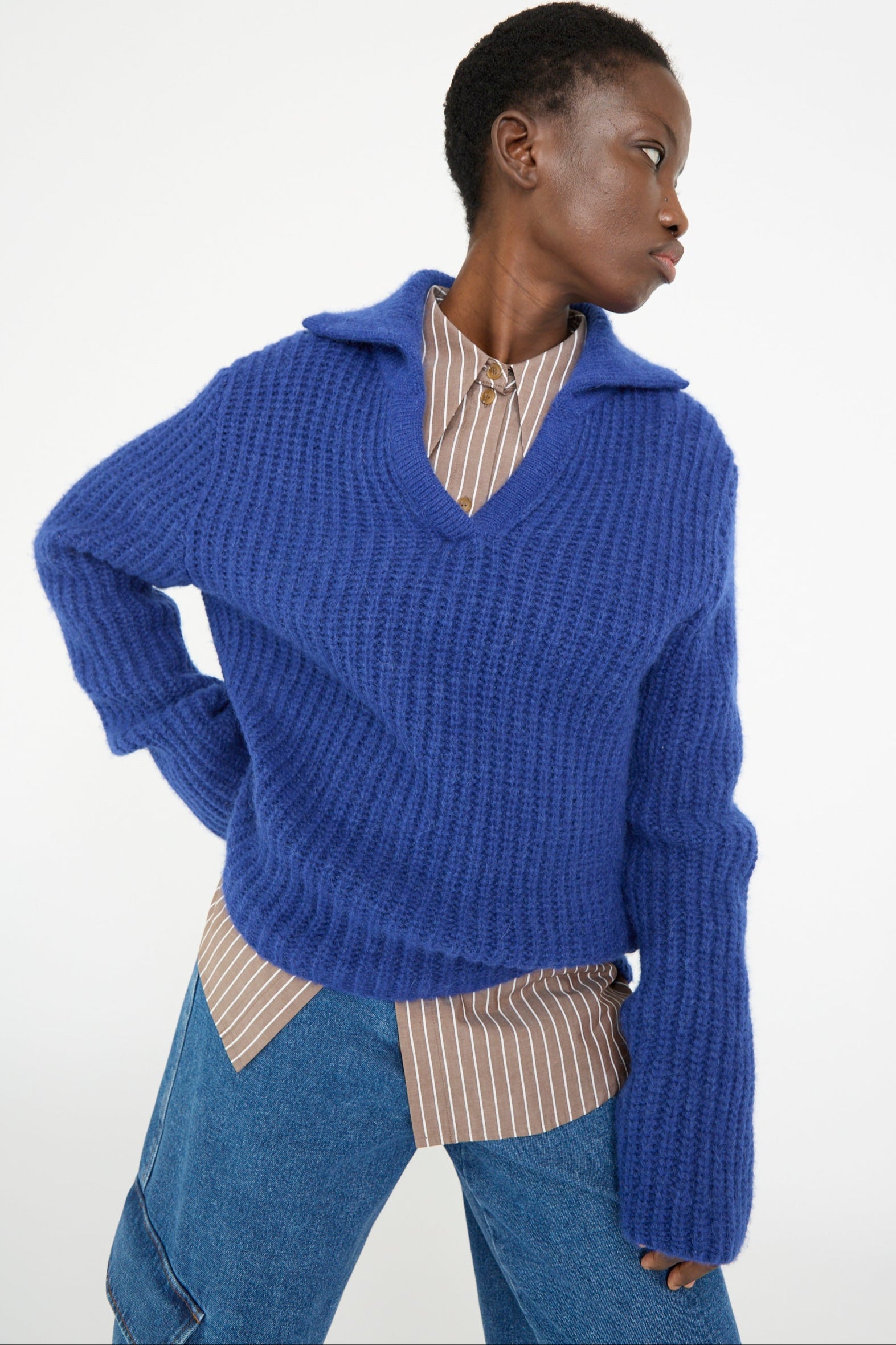 A person wears the Rejina Pyo Chunky Alpaca Alison Sweater in electric blue over a brown and white striped shirt, paired with blue pants, while looking to the side against a plain backdrop.