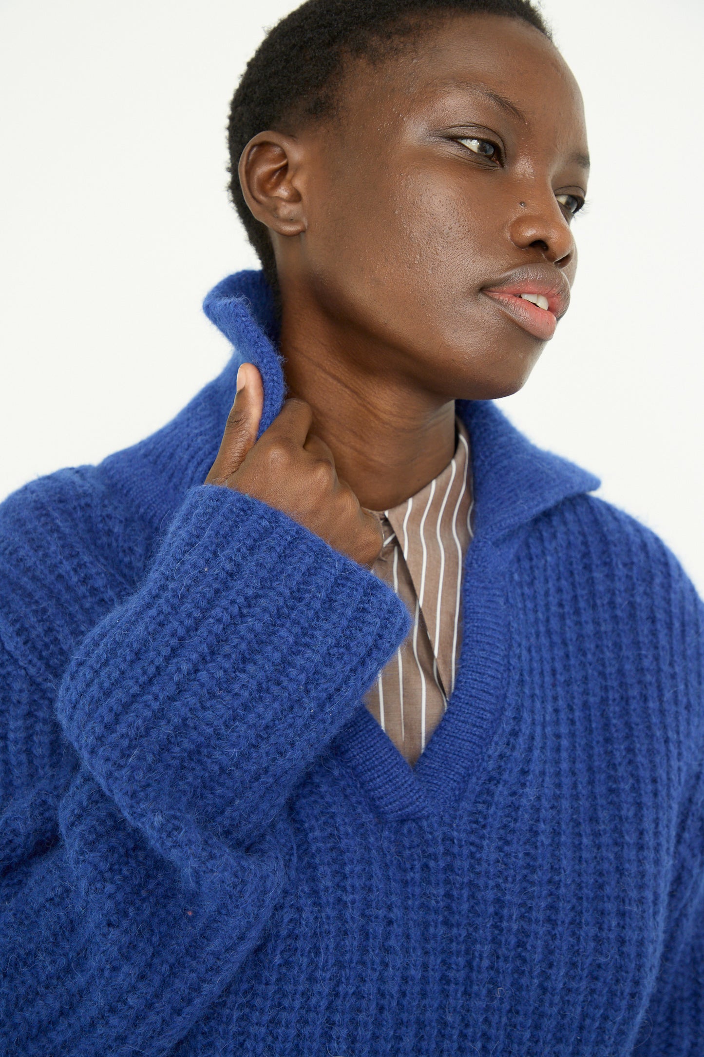 A person in a Rejina Pyo Chunky Alpaca Alison Sweater in Electric Blue and a striped shirt gazes sideways, adjusting the collar.