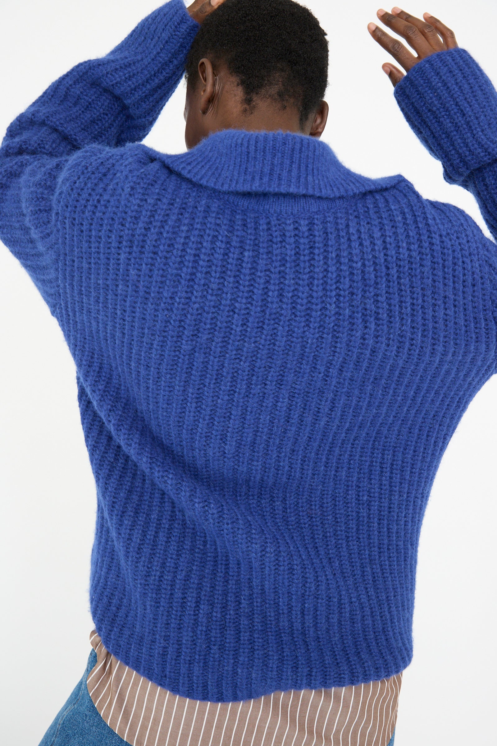 A person wearing the Rejina Pyo Chunky Alpaca Alison Sweater in Electric Blue, layered over a striped shirt, faces away with arms raised against a light background.