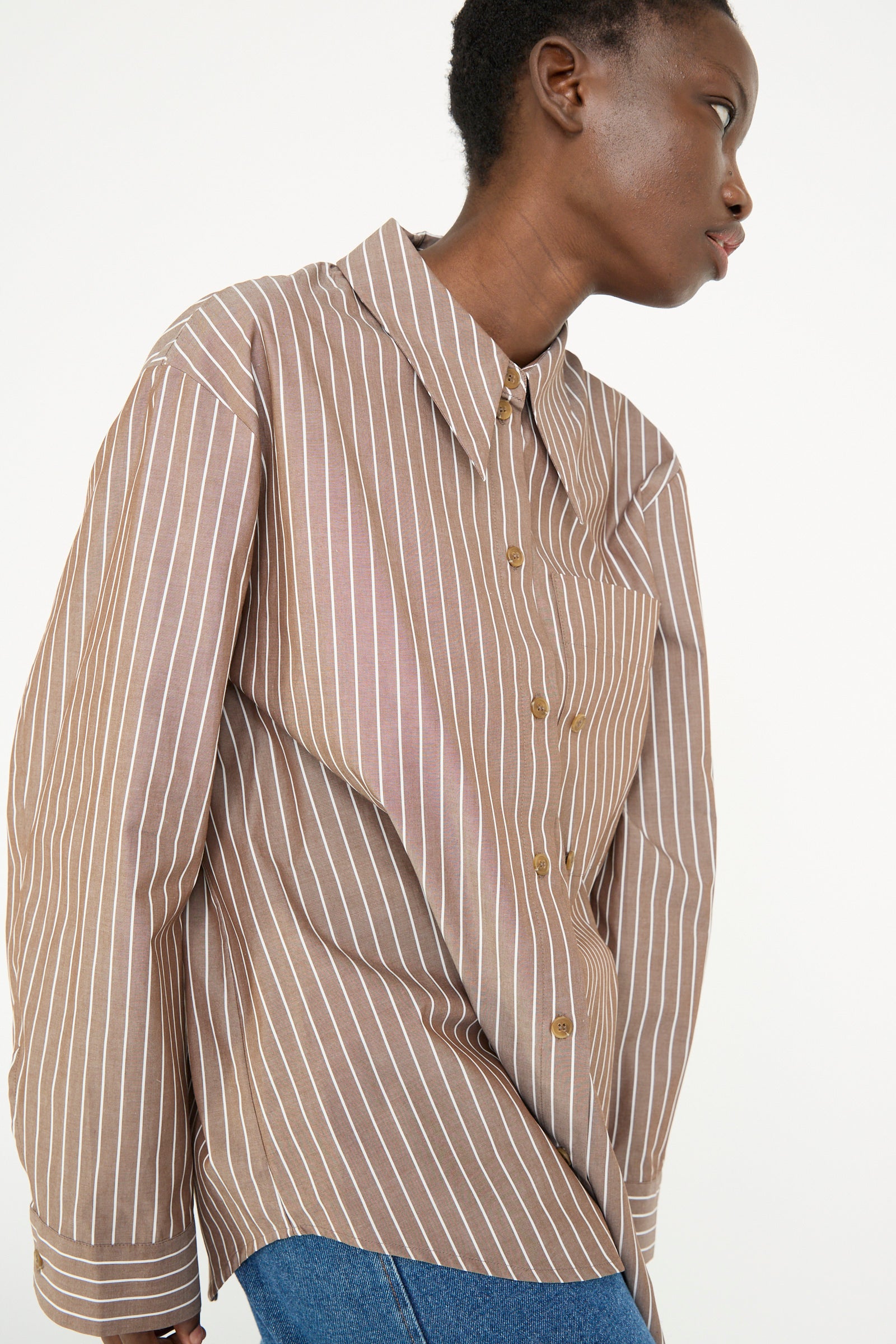 A person is wearing the Cotton Stripe Ellis Shirt in Brown by Rejina Pyo, which features a brown and white striped cotton design with an oversized fit, paired with blue jeans against a plain background.