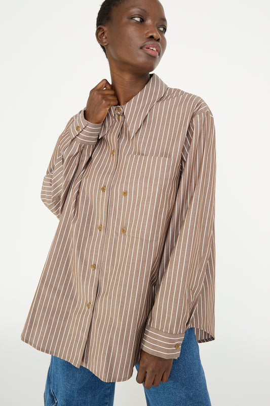 A person wearing a Rejina Pyo Cotton Stripe Ellis Shirt in Brown with blue jeans, looking to the side.